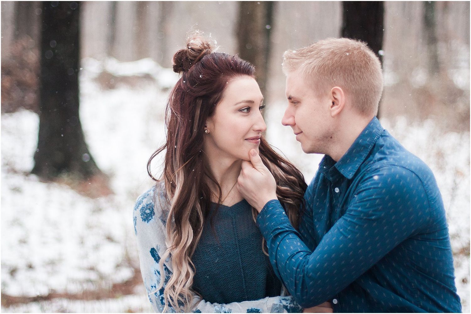 Romantic winter engagement photo of couple in snowy forest setting.
