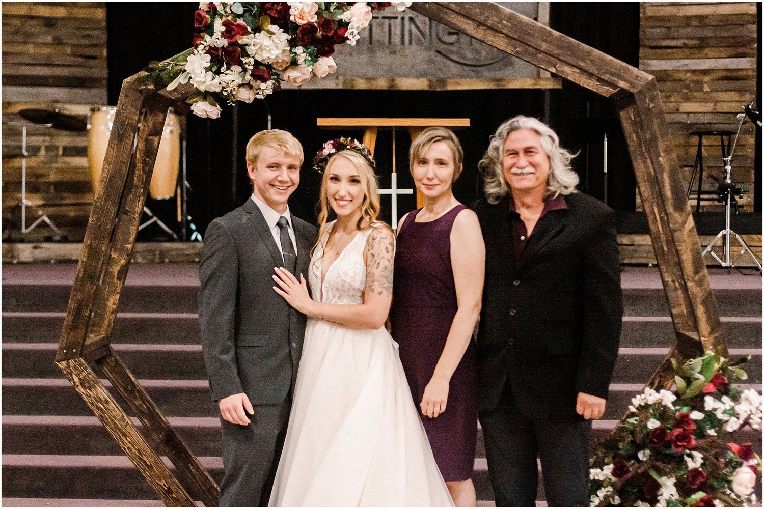 Wedding party posing under rustic wooden arch decorated with burgundy and white flowers.