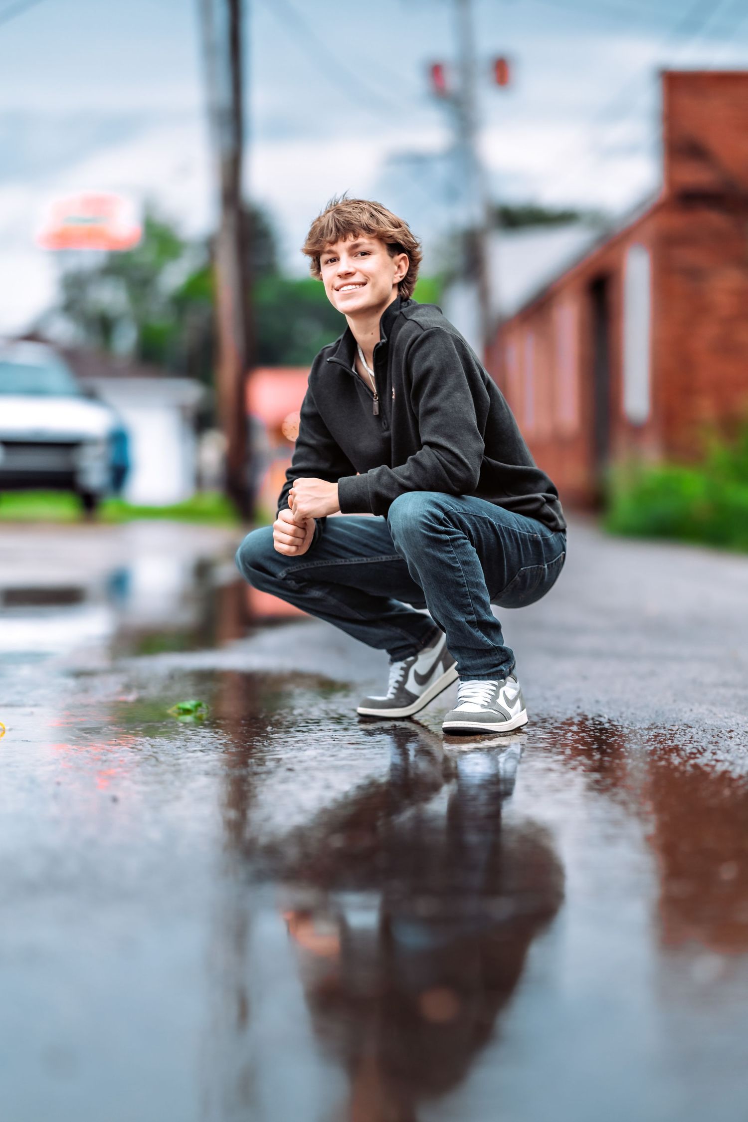 Person in black jacket crouches near a puddle reflecting the urban surroundings.