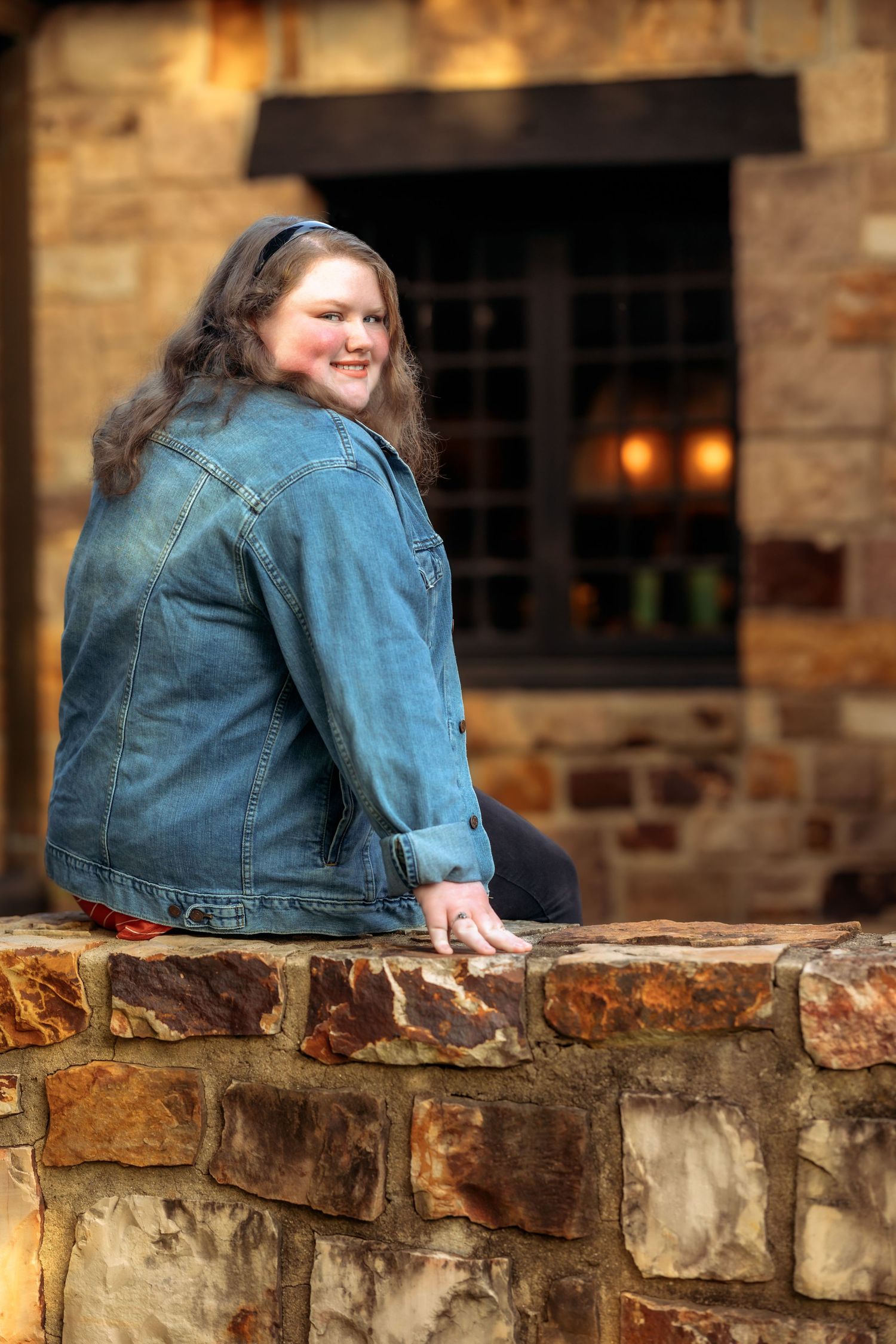 Person in denim jacket poses on rustic stone wall at sunset.