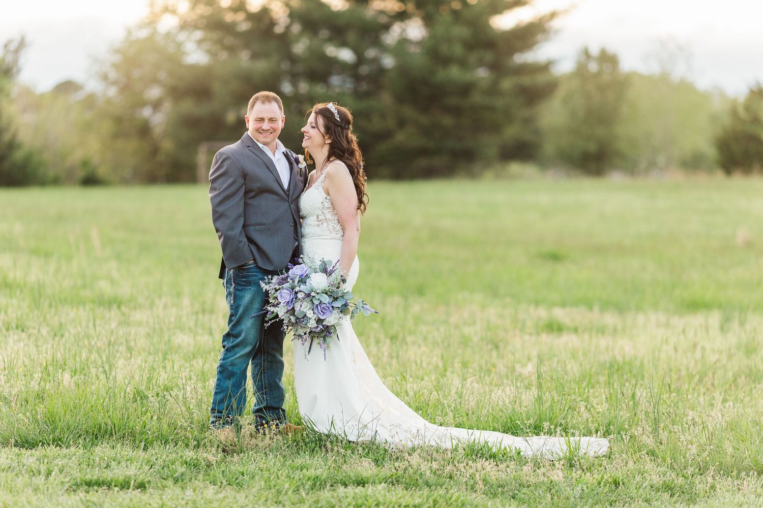 A couple stands together in a grassy field with the bride holding a blue bouquet during their outdoor wedding.