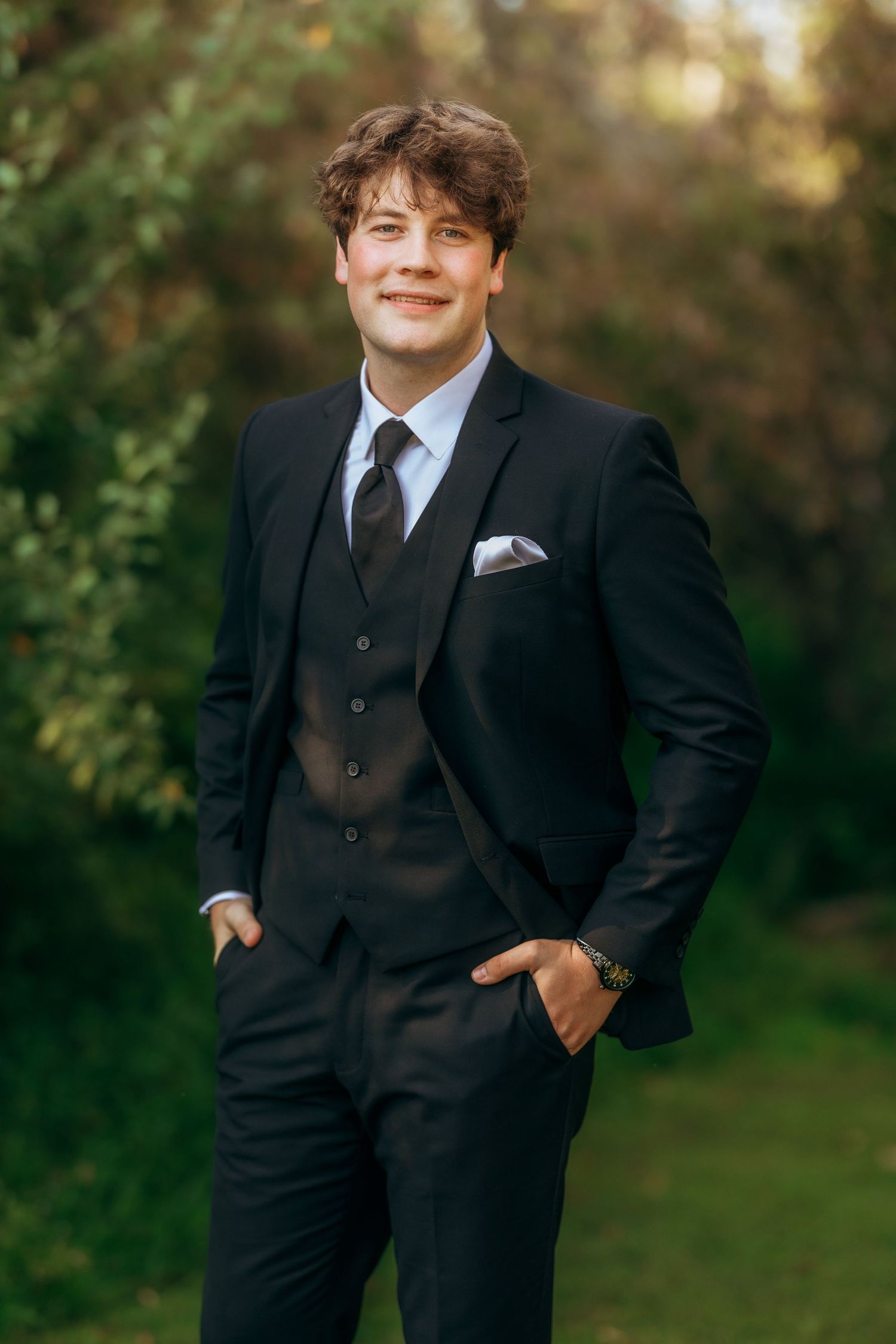Groomsman in classic black three-piece suit poses in outdoor garden setting.
