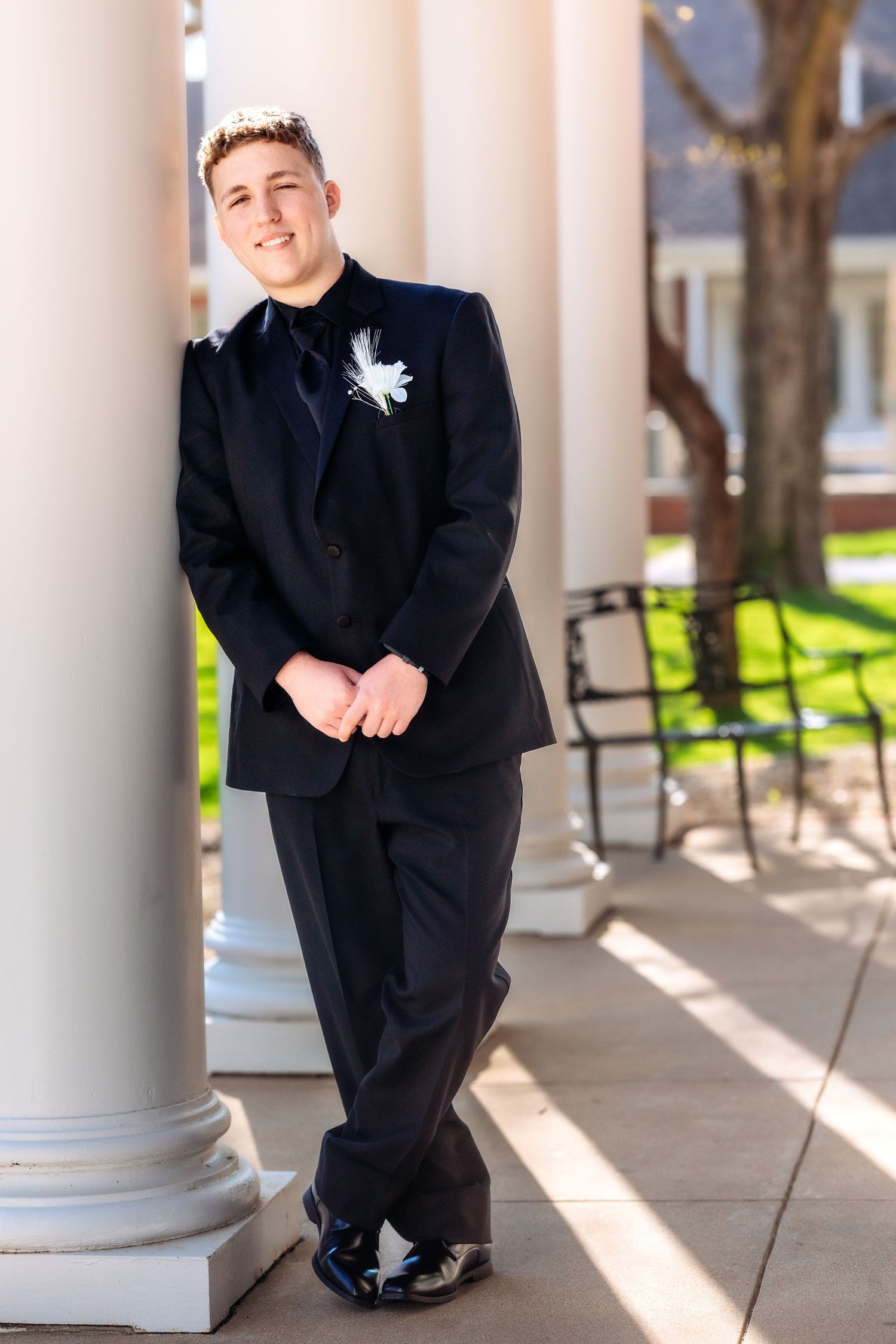 A formal portrait session showing someone in a black tuxedo posing against white columns in natural sunlight.