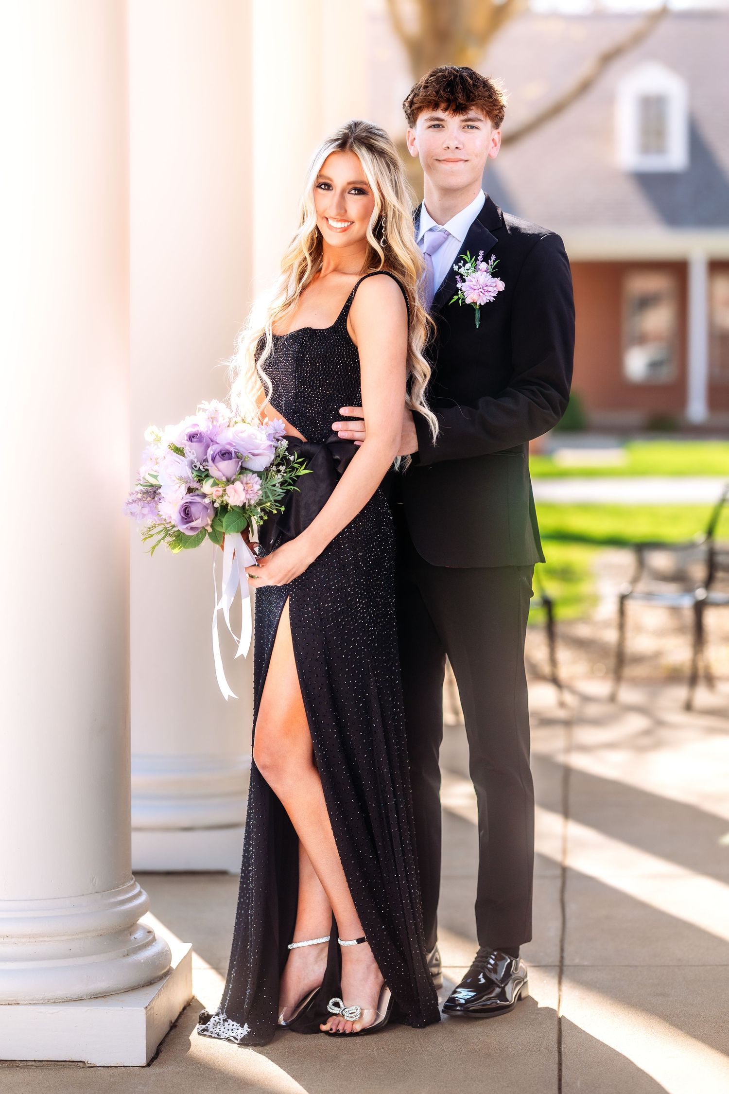 A couple in formal attire poses together near white columns holding purple flowers at a special event.