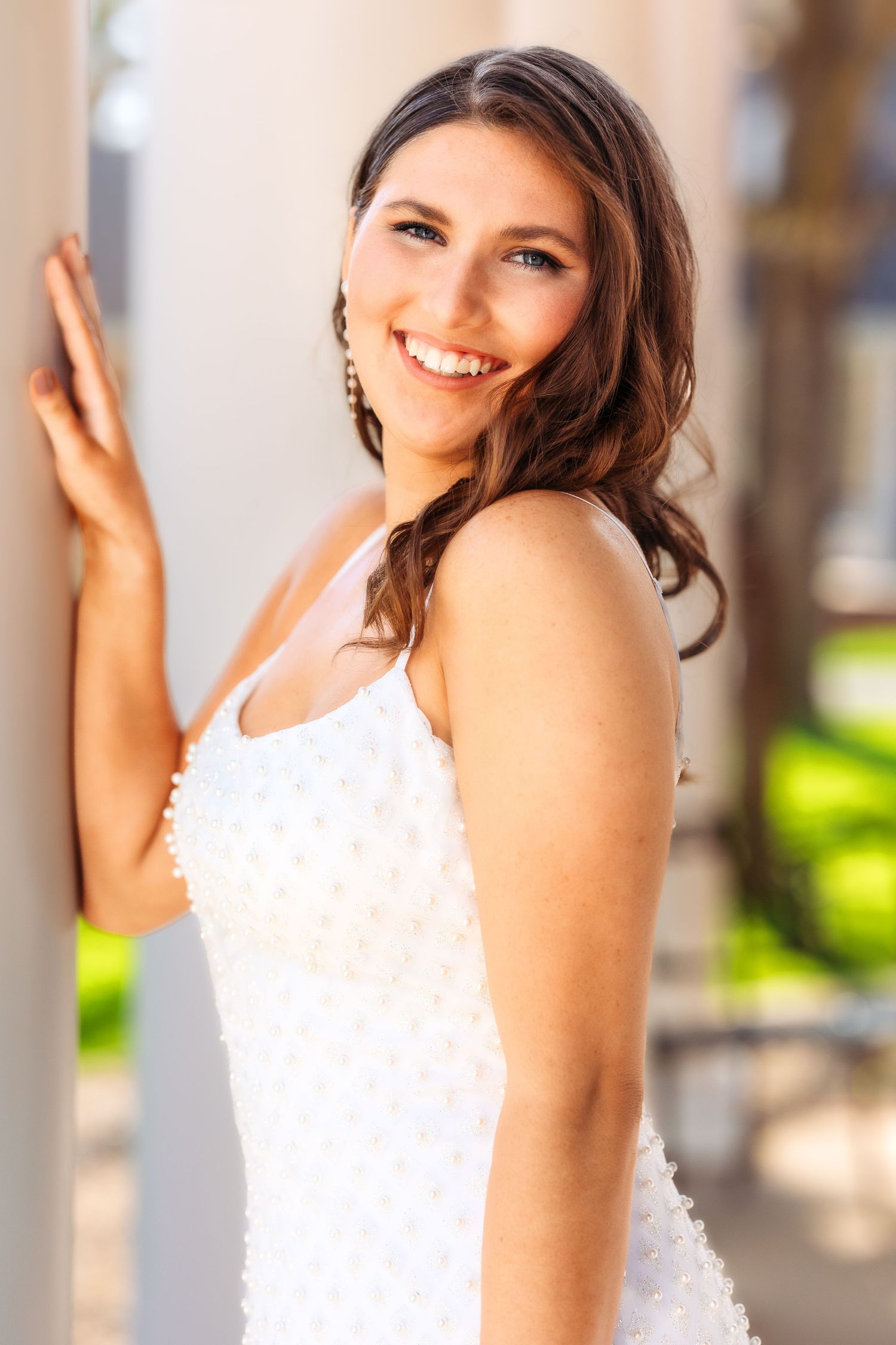 A bride in a white wedding dress poses elegantly against white columns with natural lighting and greenery in background.