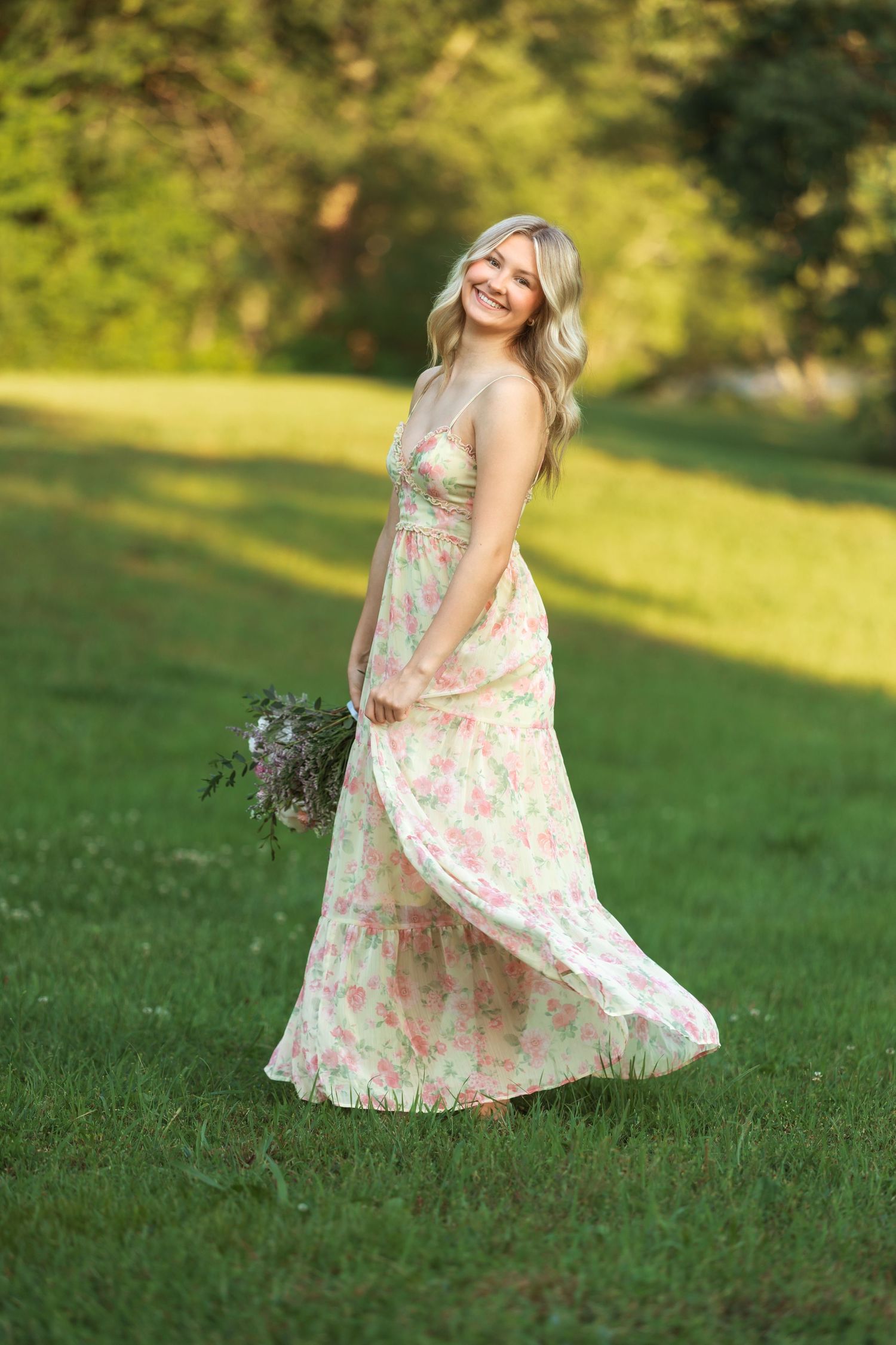 Smiling blonde woman in floral maxi dress holding bouquet, twirling in green park on sunny day.