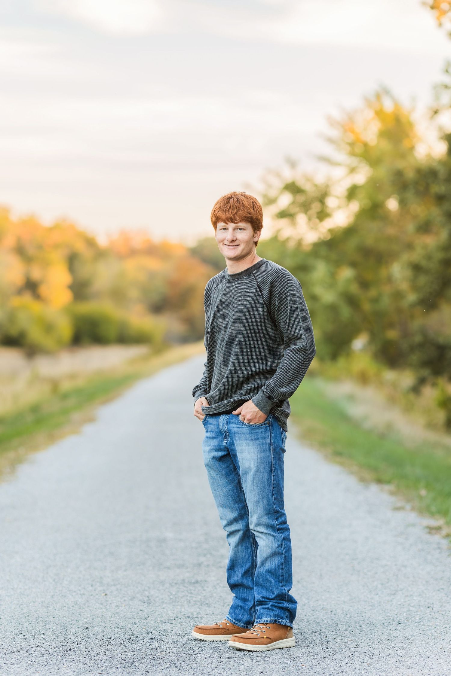 A casual portrait of someone in a grey sweater standing on a rural path in autumn.