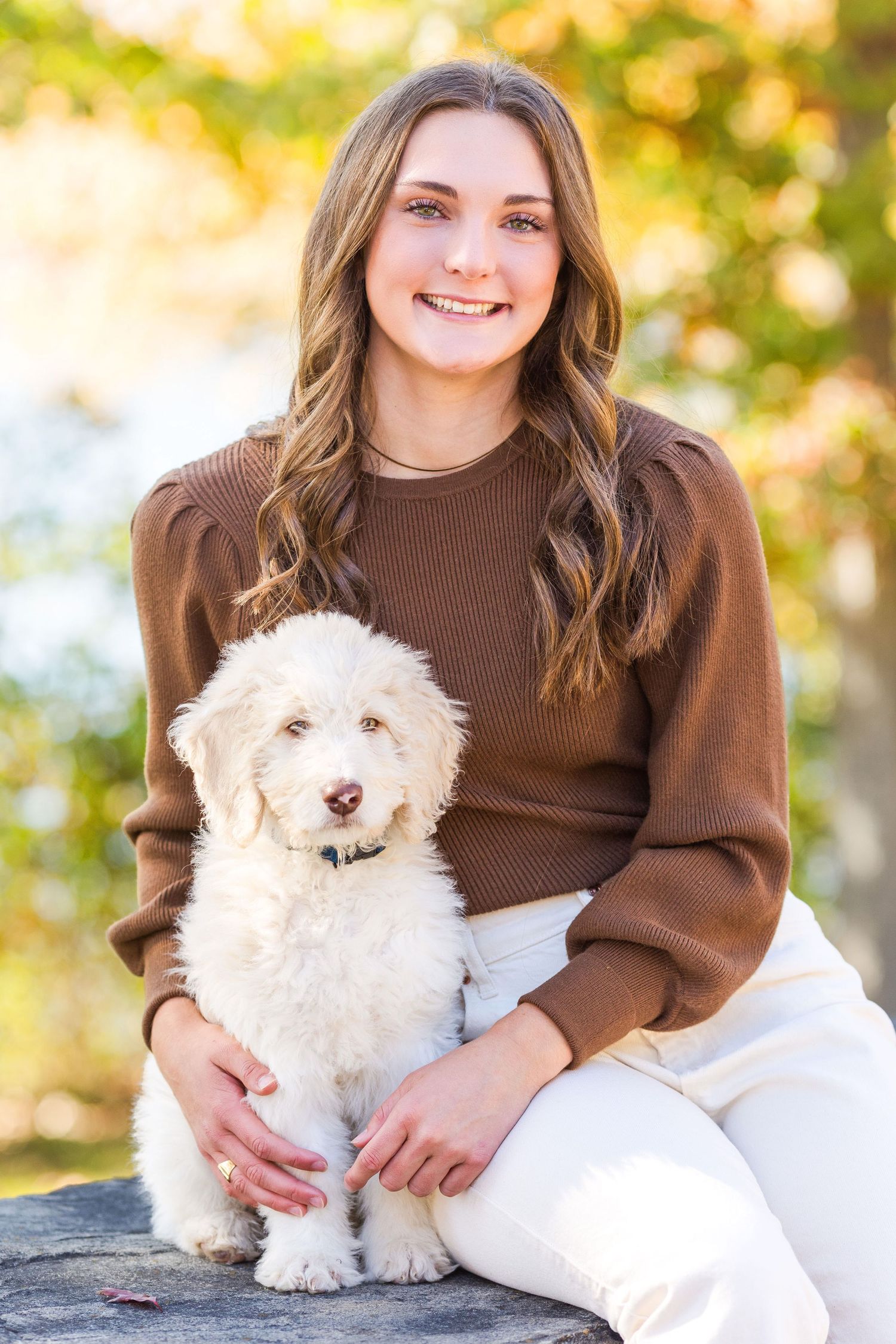 A person in a brown sweater holds a small white fluffy dog while sitting outdoors in autumn.