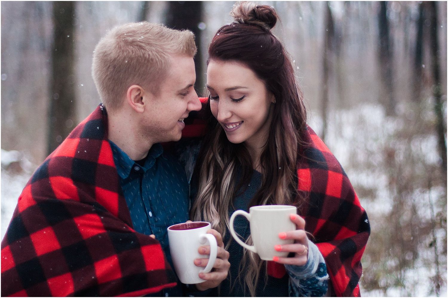 A couple shares hot drinks wrapped in a red buffalo plaid blanket during a winter photoshoot.