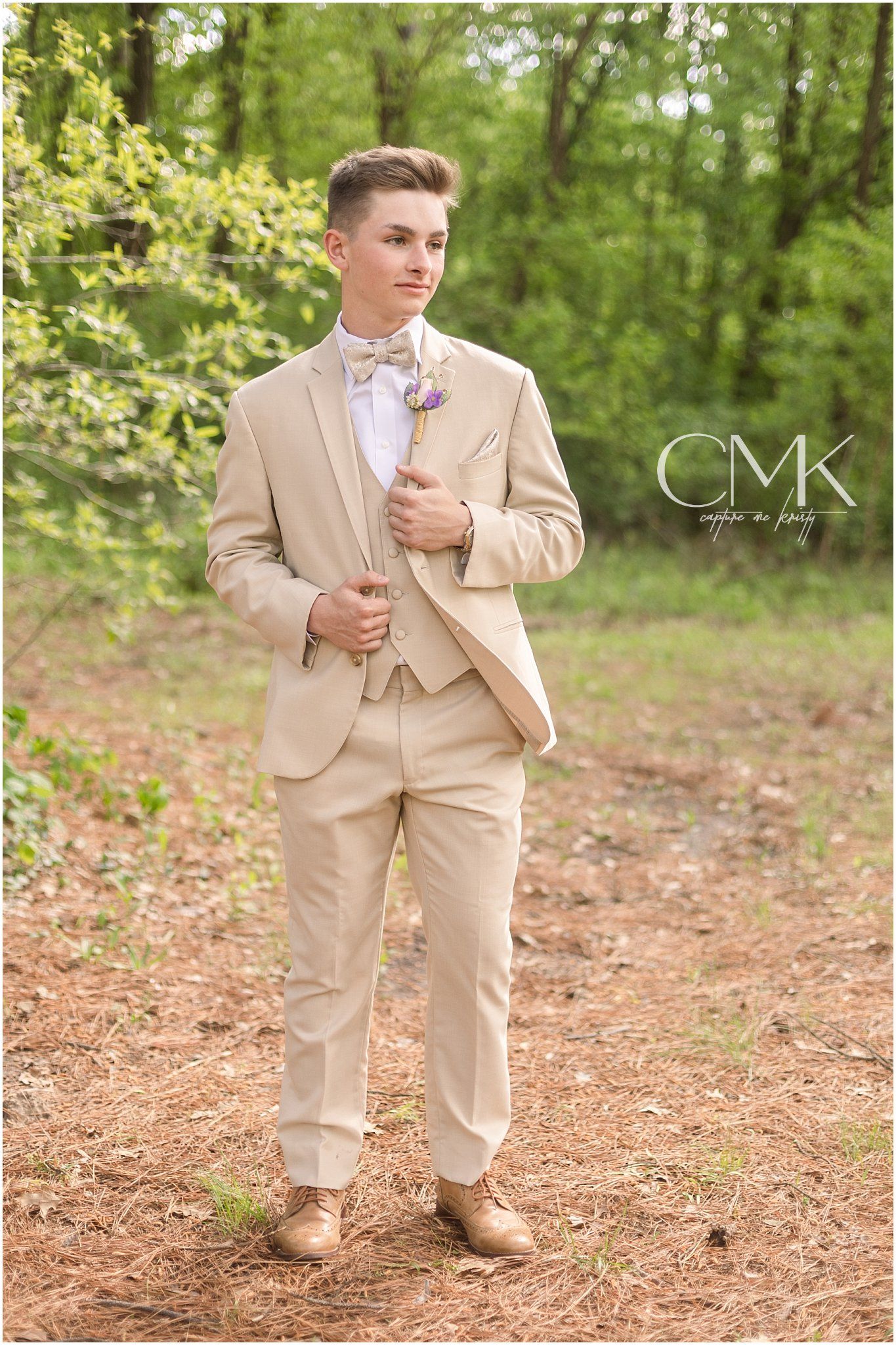 A formal portrait featuring a beige suit and pink bow tie in a wooded setting.