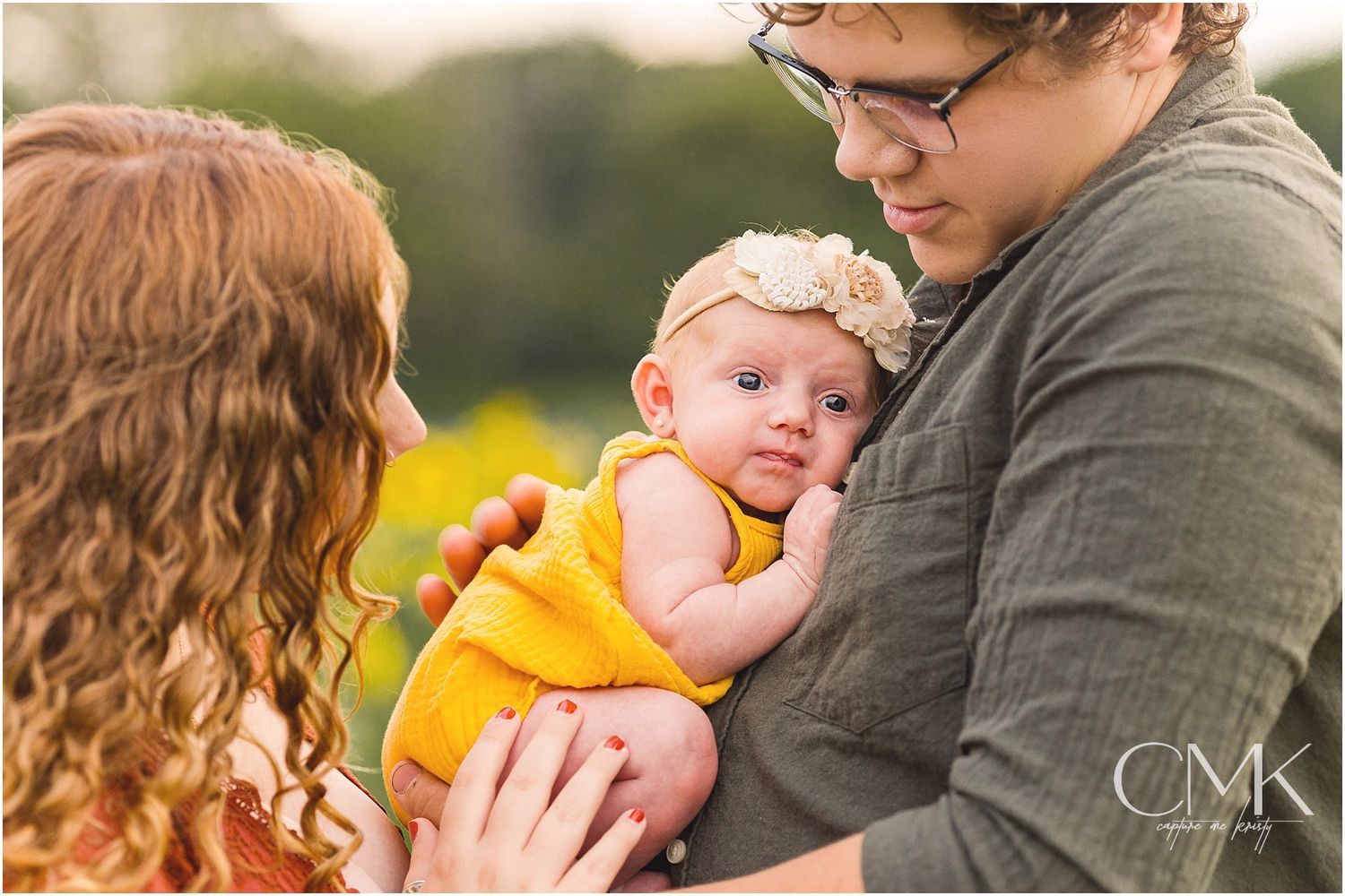 Close-up shot of parents cradling their baby in yellow against a blurred nature background.