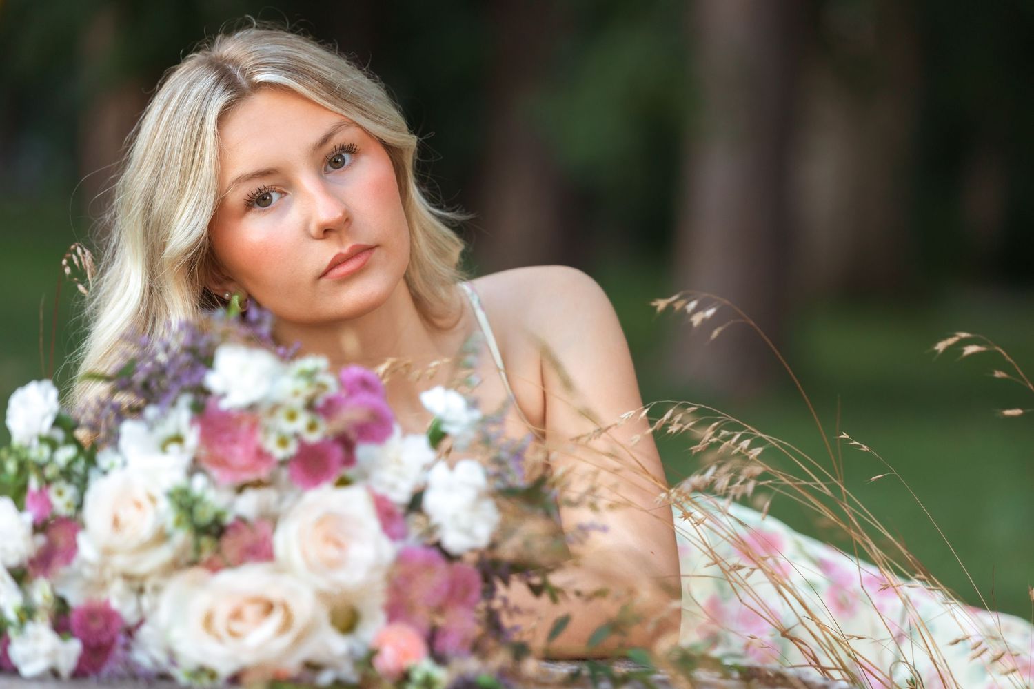 Blonde bride lying in grass with a lush bouquet of pink and white roses in a romantic outdoor setting.