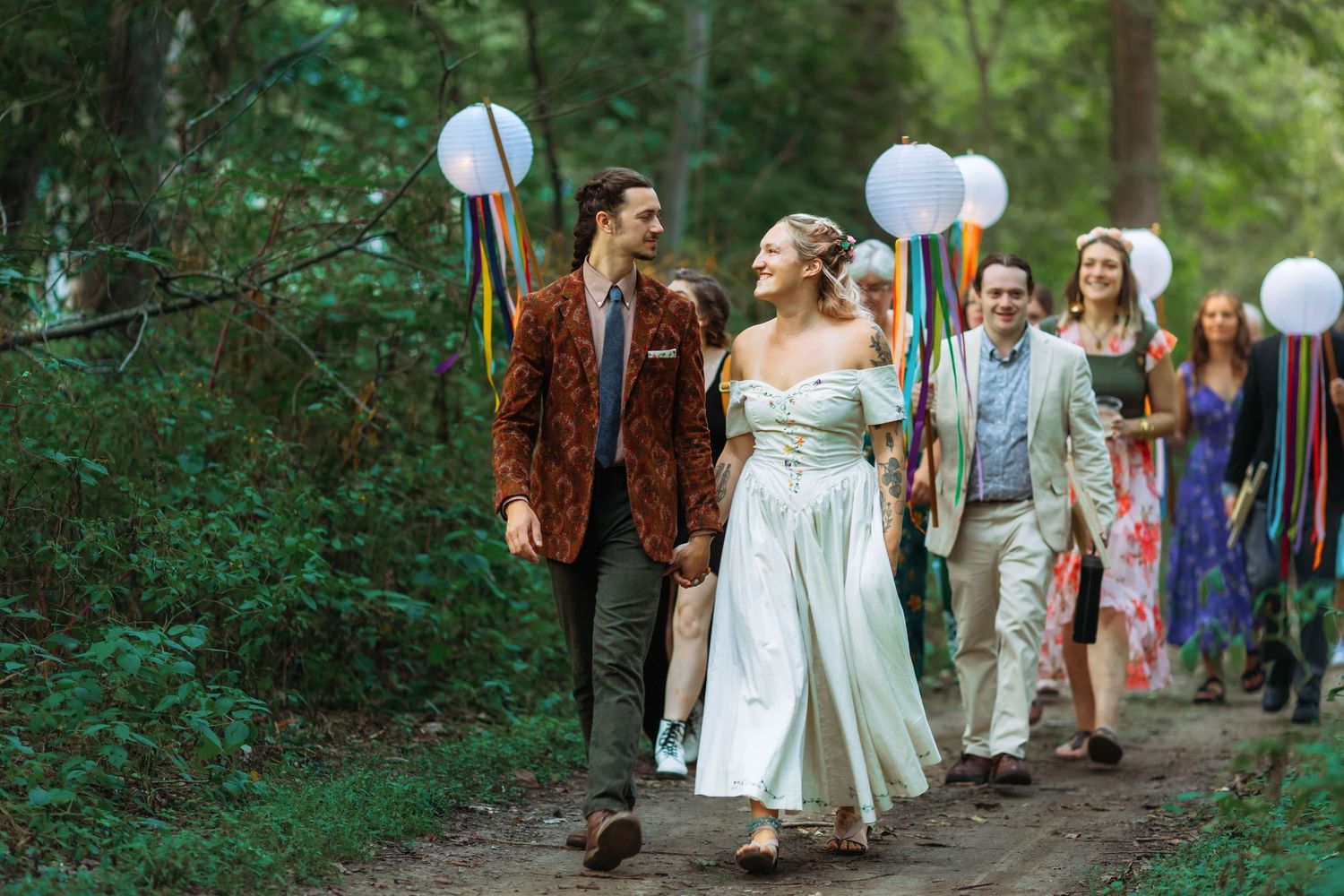Wedding guests walking through wooded path with white lanterns.