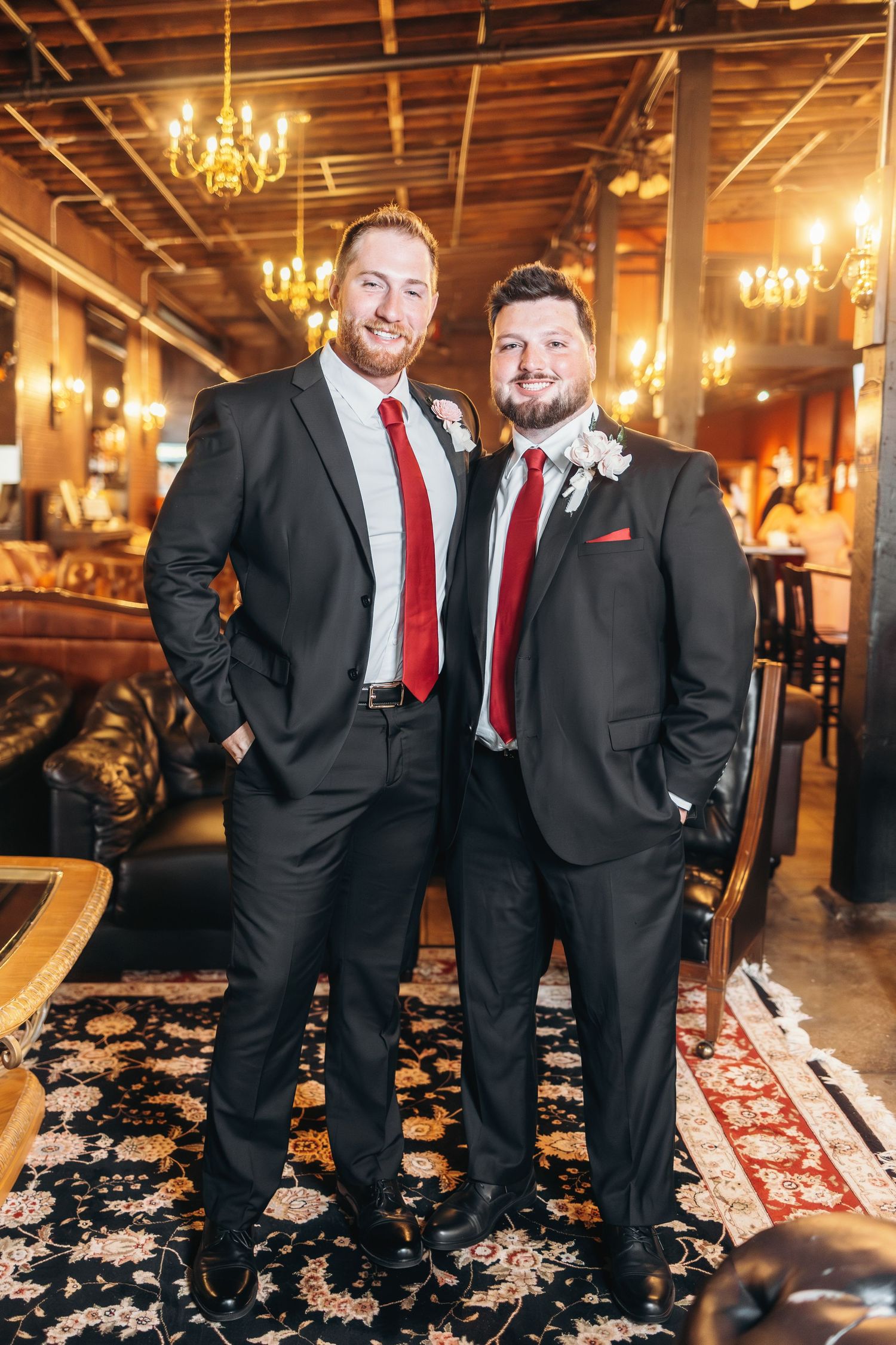 Groomsmen in black suits with red accents pose on oriental rugs under warm lighting.