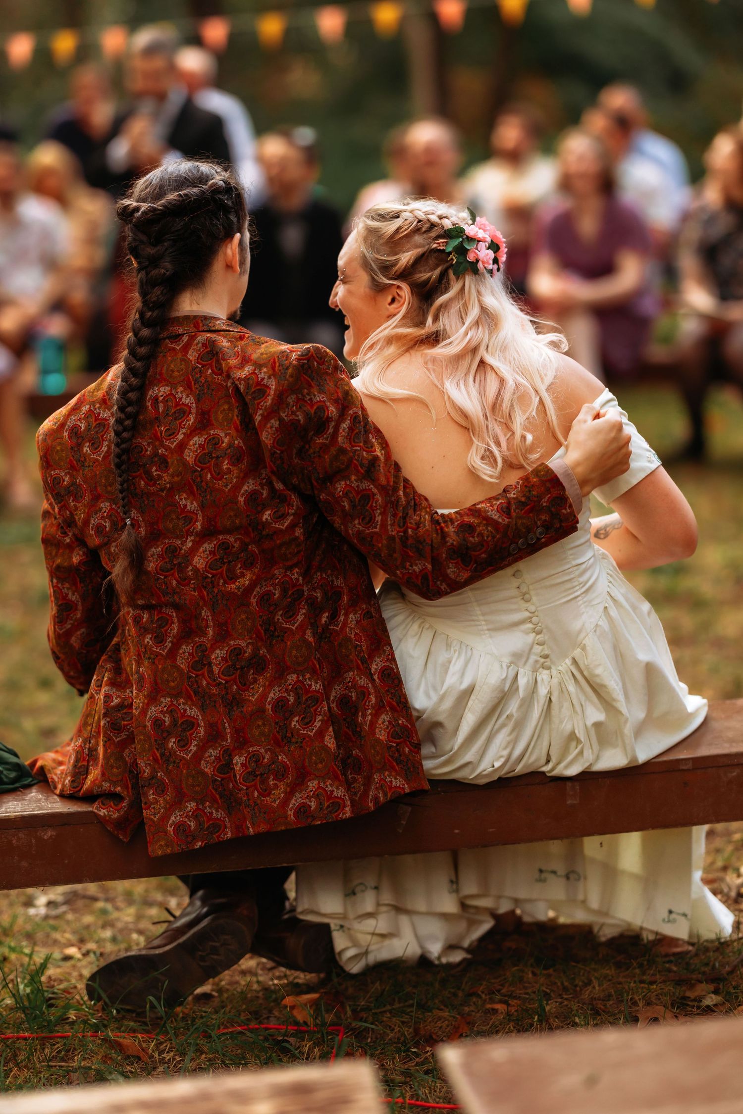 A couple sits on a bench at an outdoor wedding, with the bride in white and partner in a burgundy patterned jacket.
