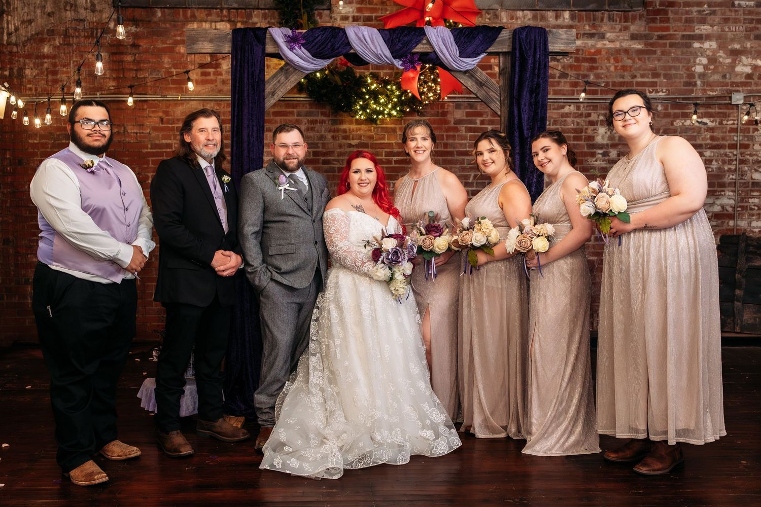 Wedding party in neutral tones poses together against rustic brick wall backdrop.