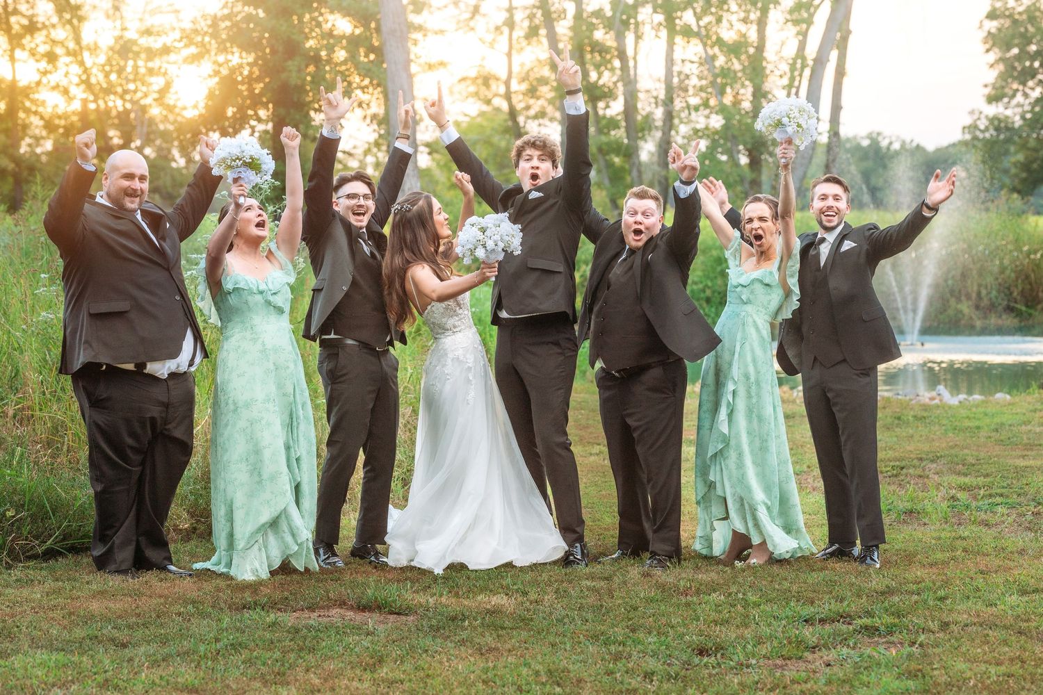 Wedding party in mint green and dark suits celebrating with raised arms at sunset.
