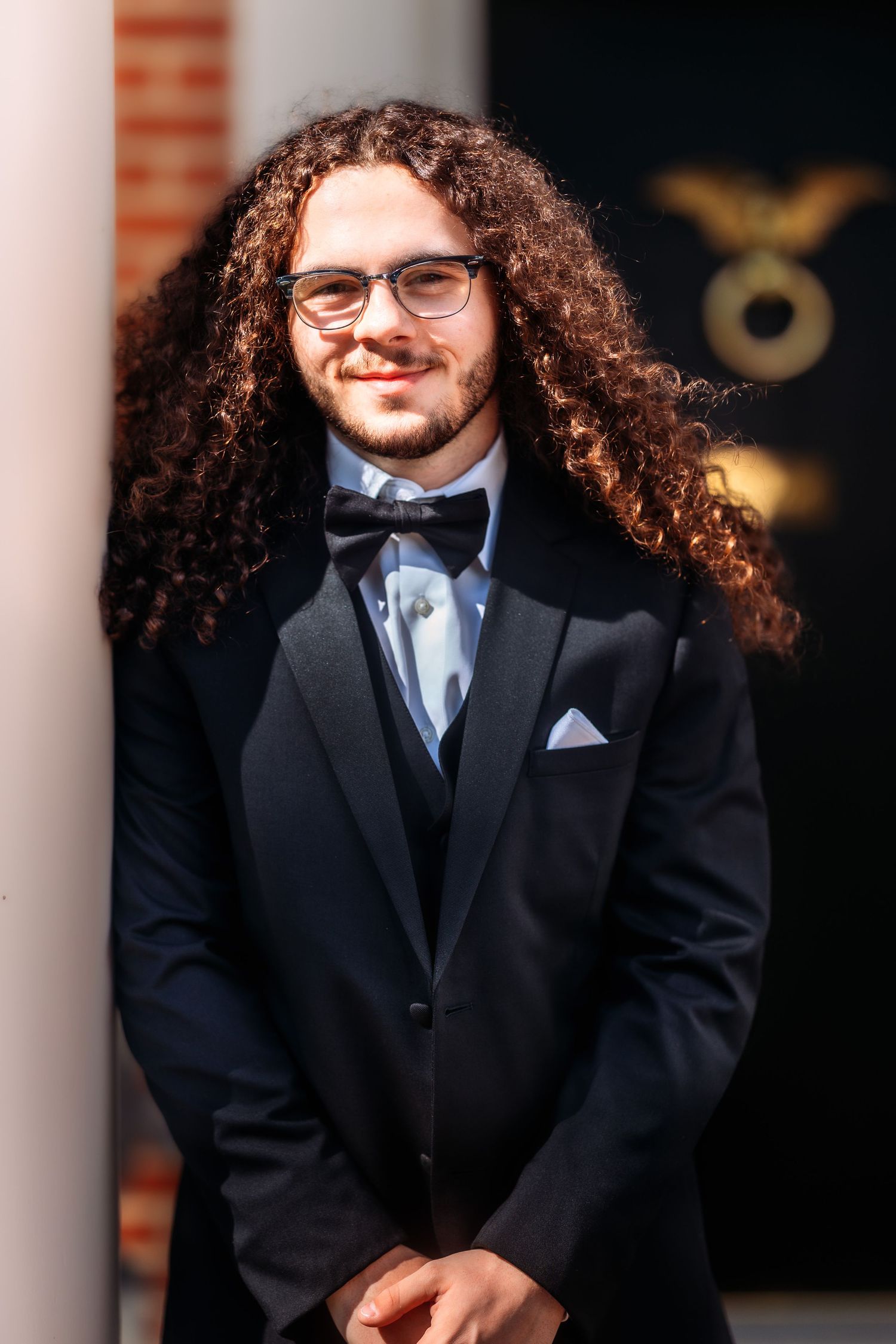 A person with long curly hair and glasses wearing a black suit and tie smiles while posing against white columns.