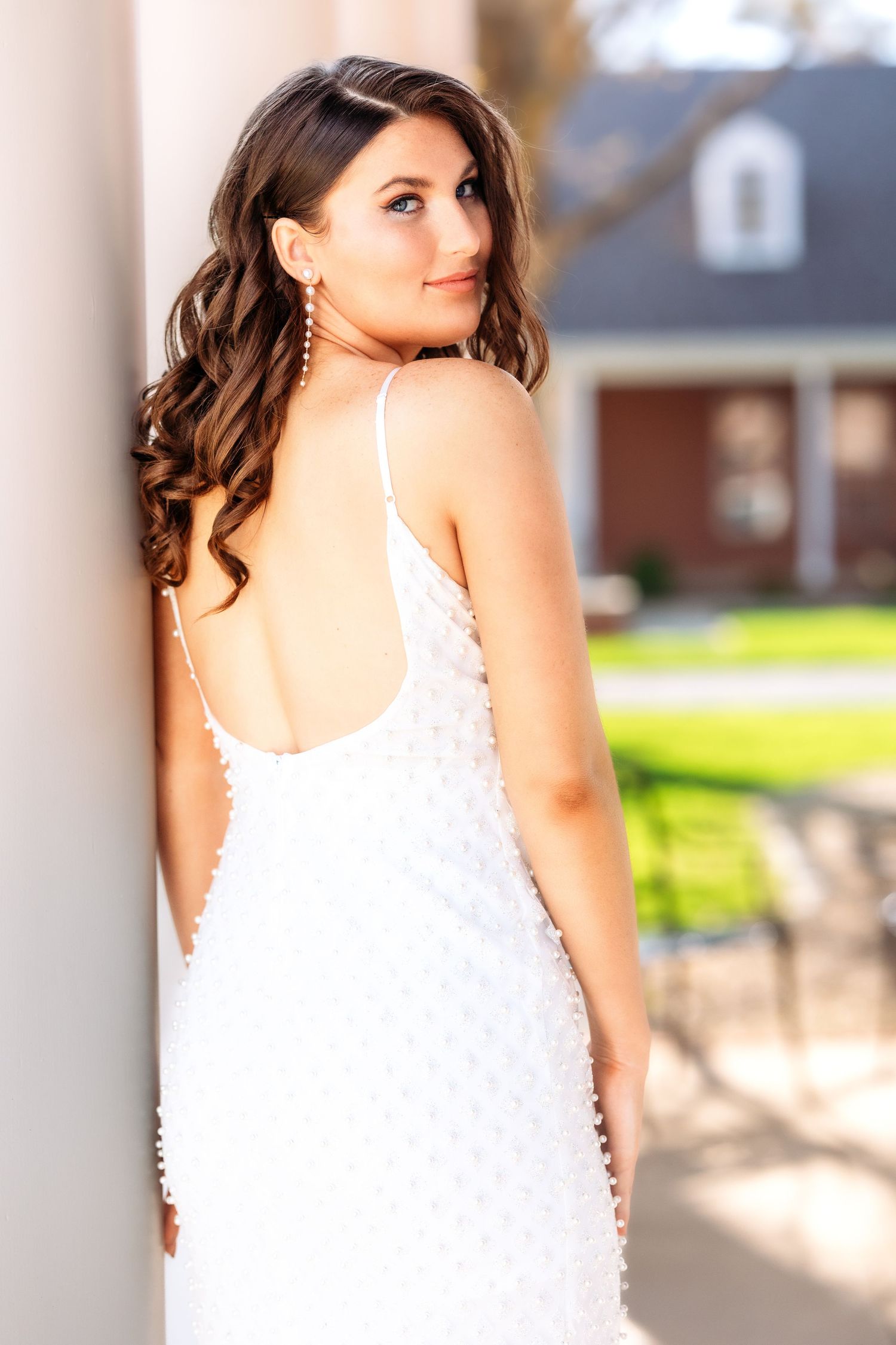A woman in a white lace dress poses elegantly against white columns outdoors on a sunny day.