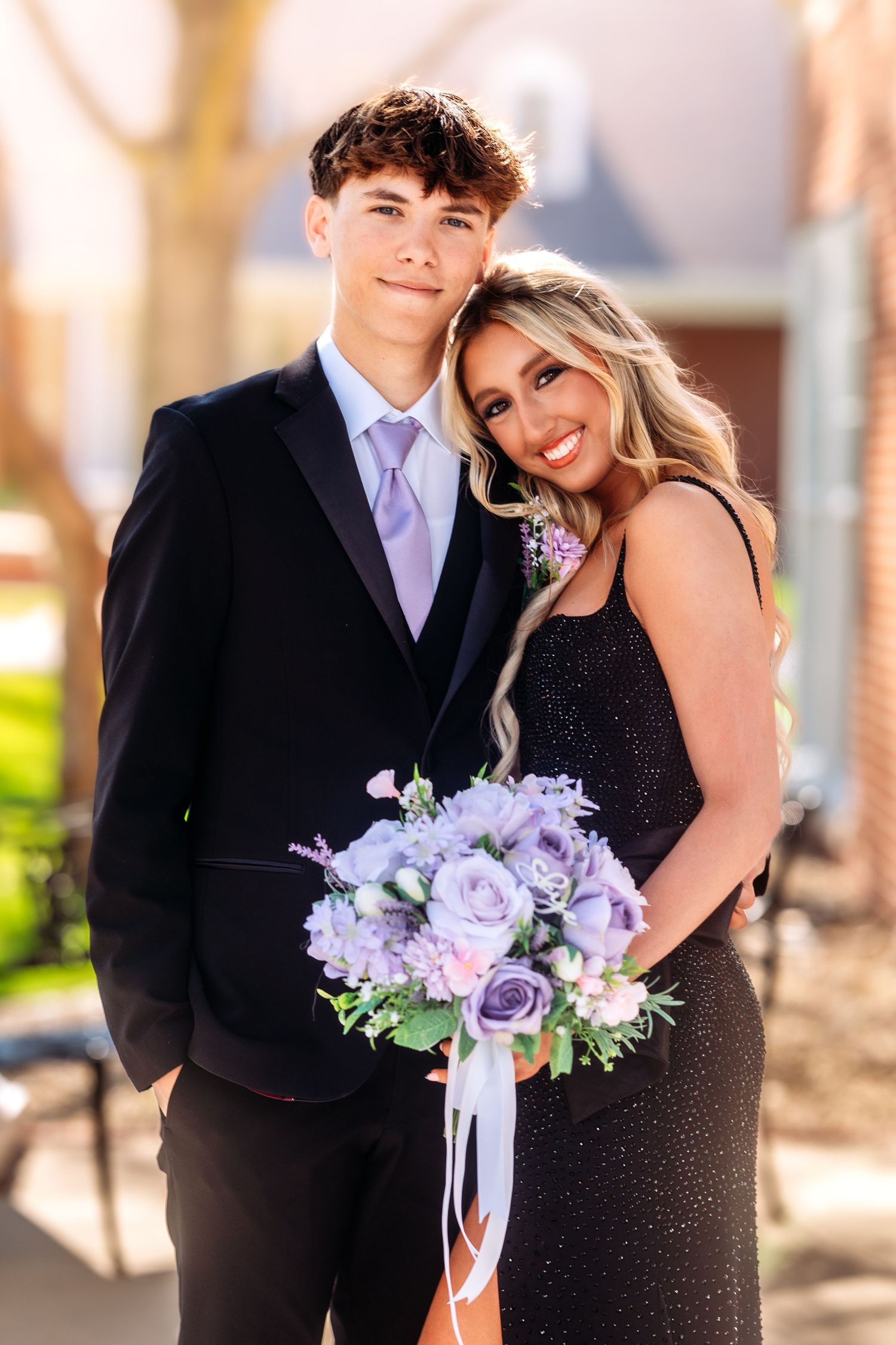Young couple in formal attire embrace at sunset with purple floral bouquet during special occasion photoshoot.