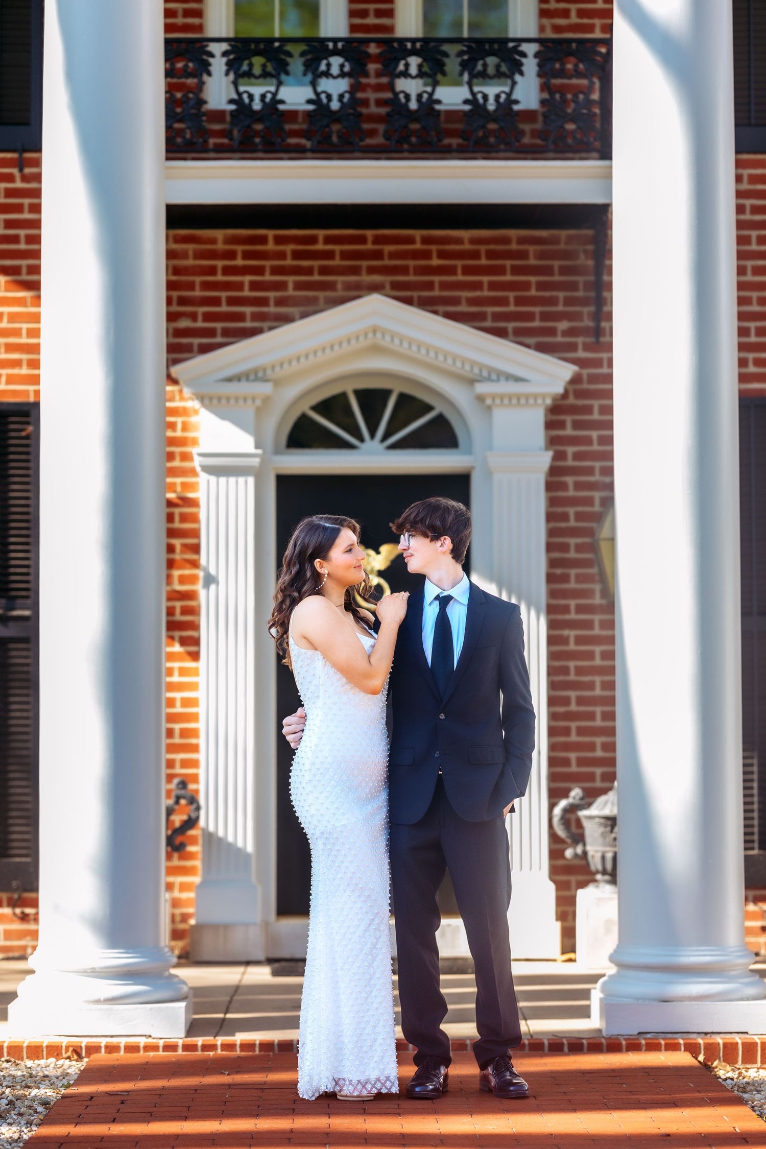 A couple in formal attire stands between white columns on the porch of a classic brick building with an arched doorway.