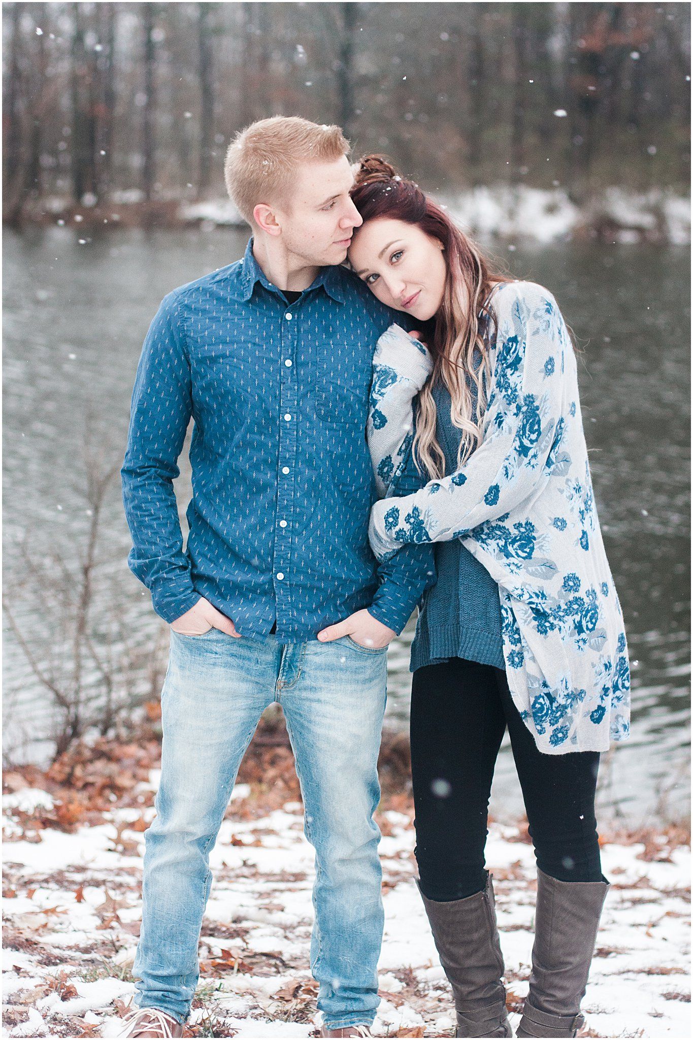 A couple in coordinating blue outfits poses by a lake during a snowy winter photoshoot.