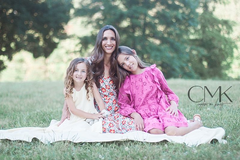 Mother and daughters sitting together on white blanket in grassy outdoor setting.