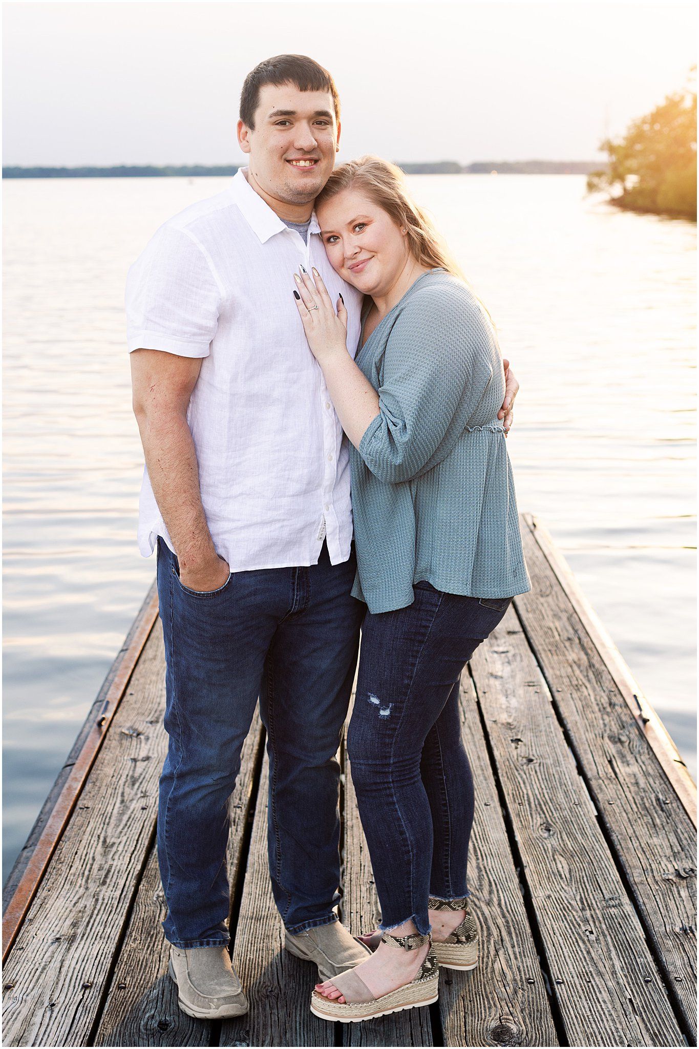 Couple stands close together on wooden dock with water and sunset in background.