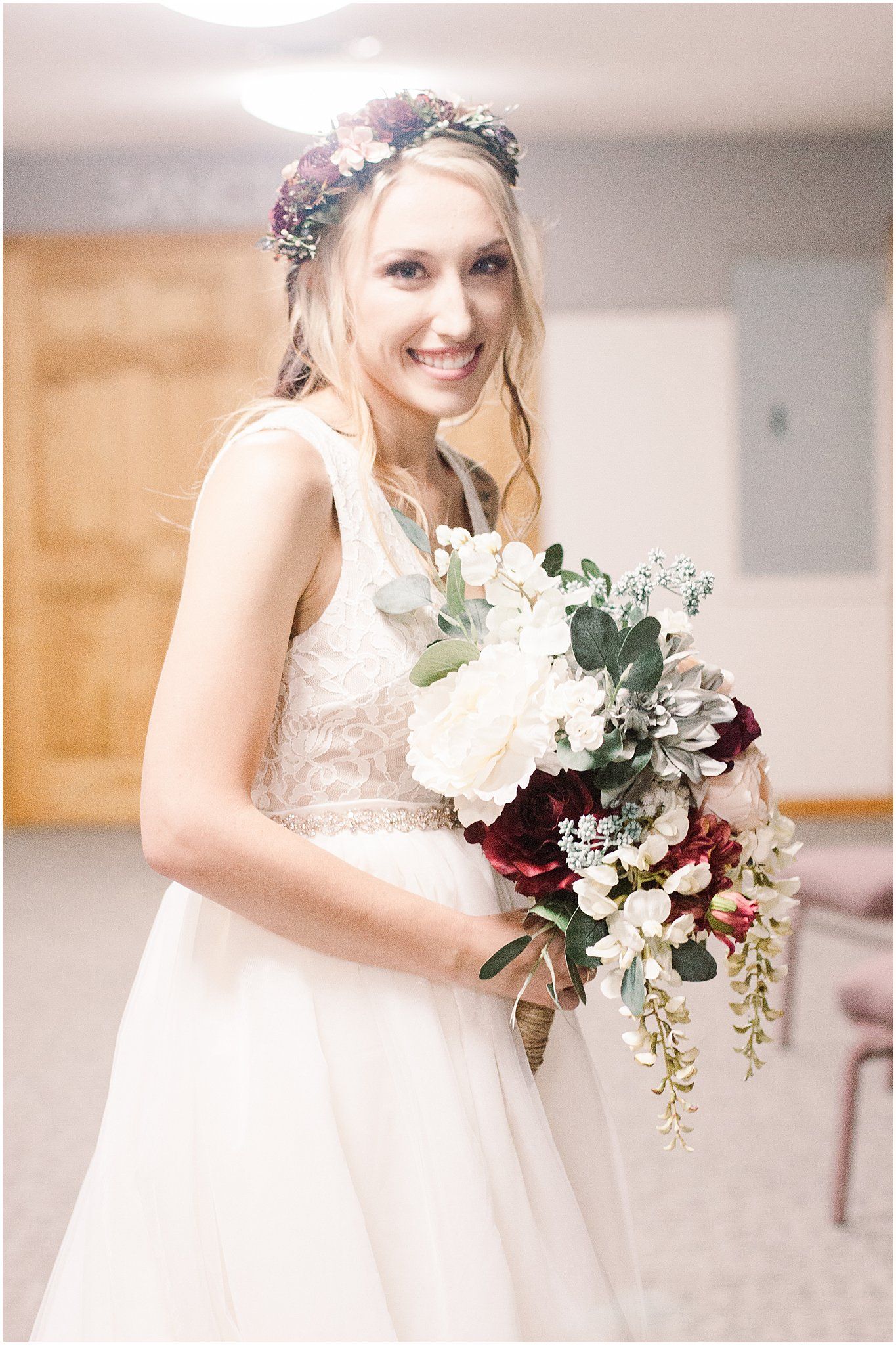 Bride wearing floral crown and lace dress holds elegant burgundy and white bouquet.