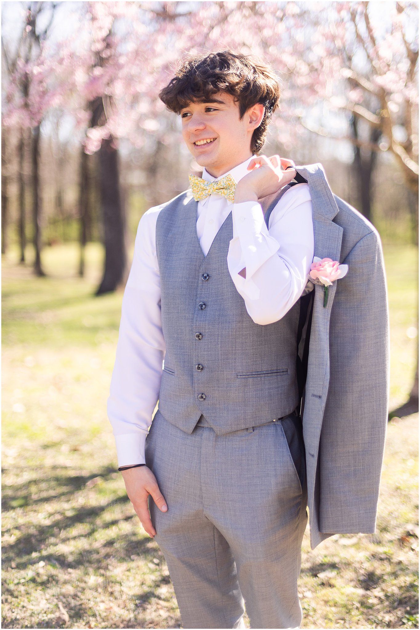 A person in a light gray suit and bowtie poses against blooming cherry blossom trees in a spring setting.