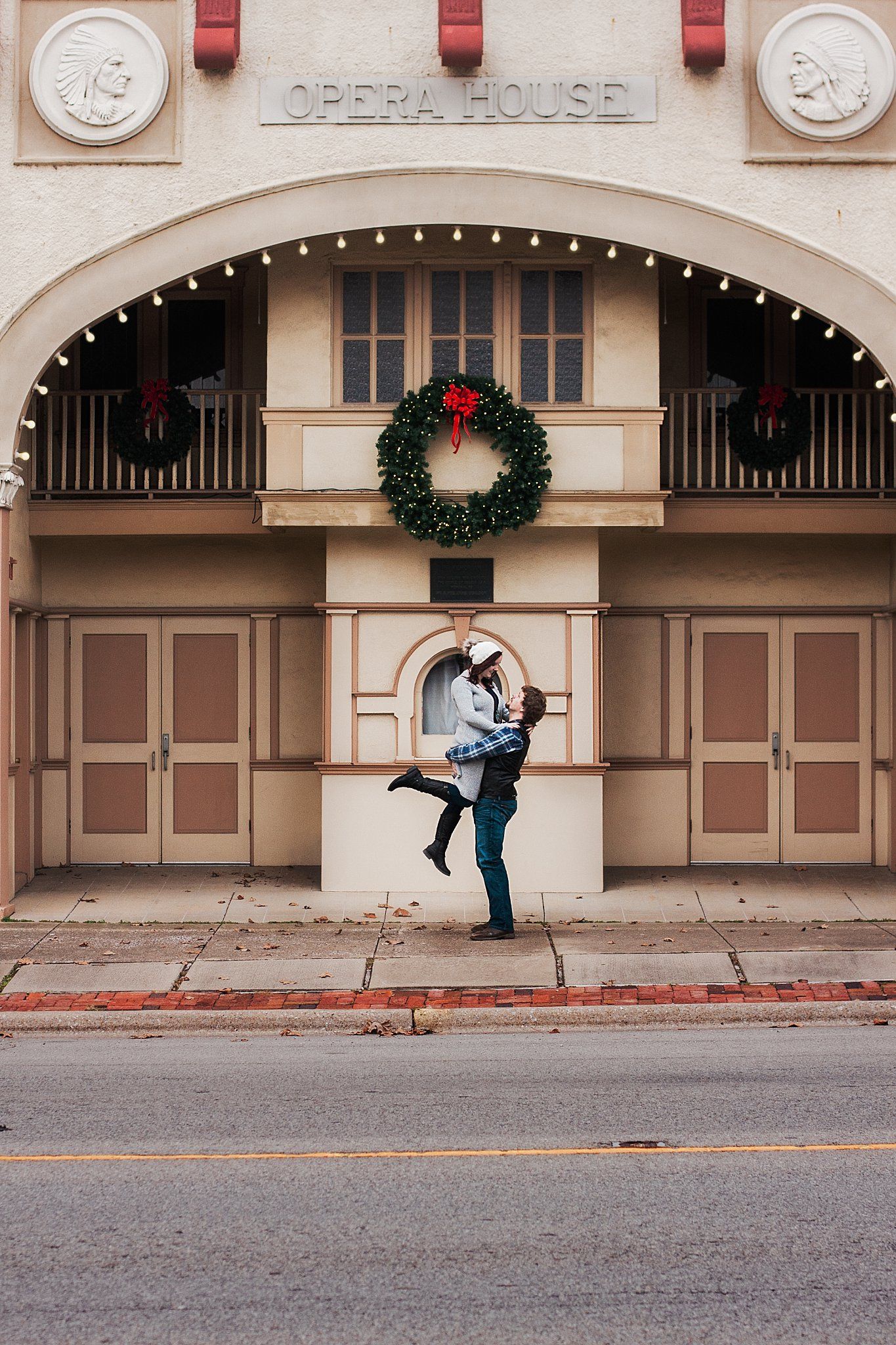 man picks up woman as she kicks her legs out while they stand infront of an old opera house