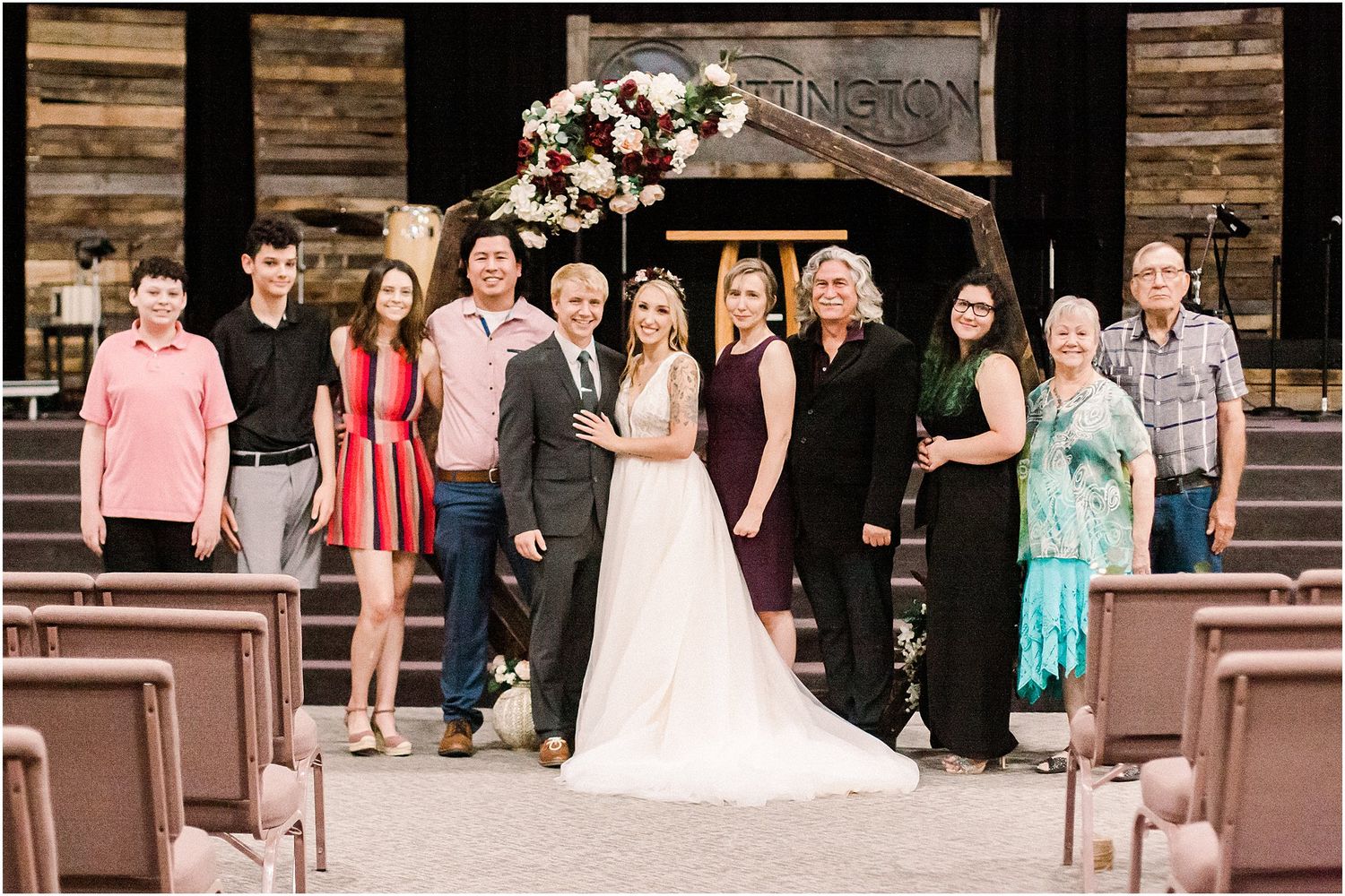 Large wedding party group photo with bridal couple centered under decorated arch.