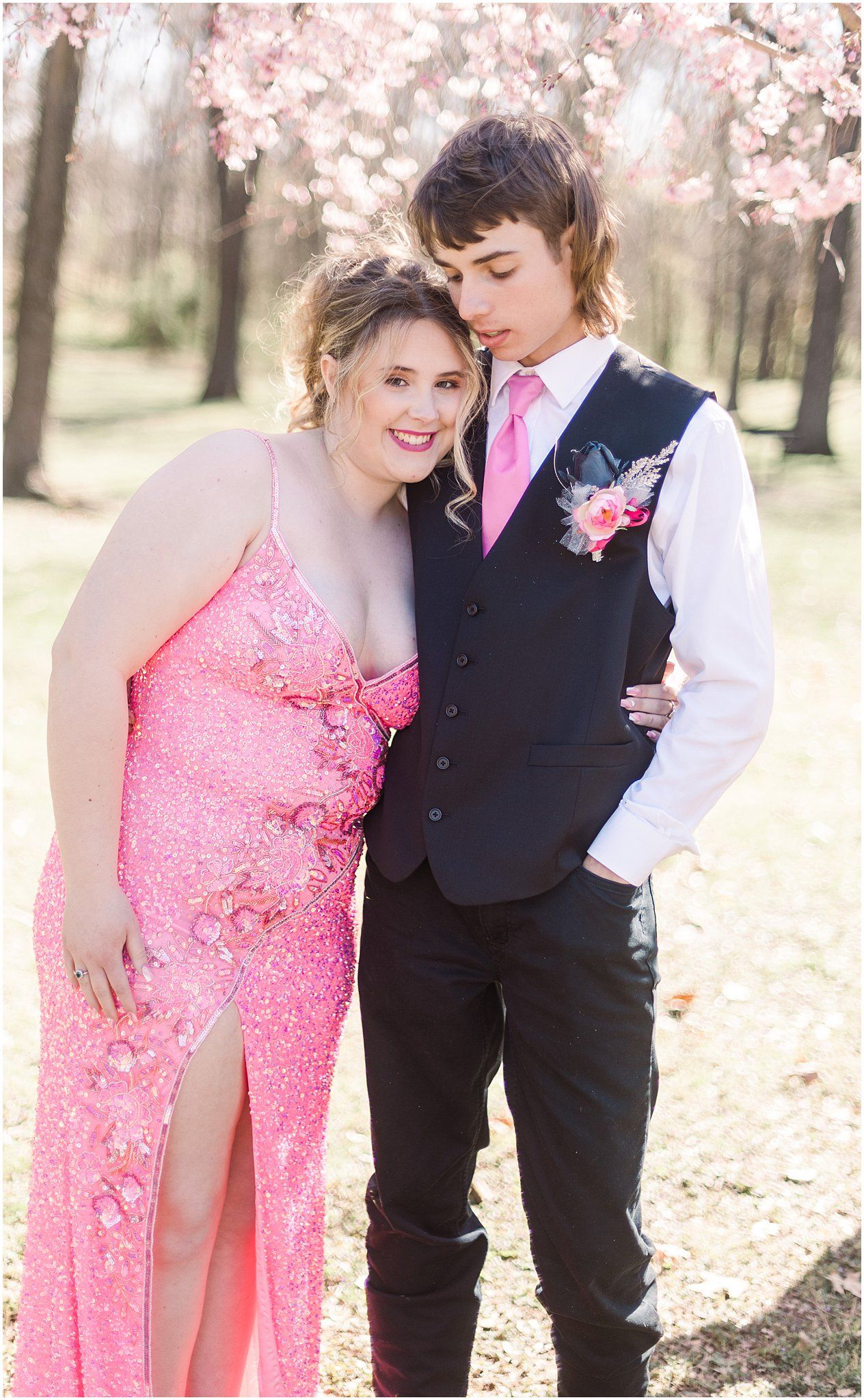 Formal couple in pink and black outfits pose close together under flowering trees.