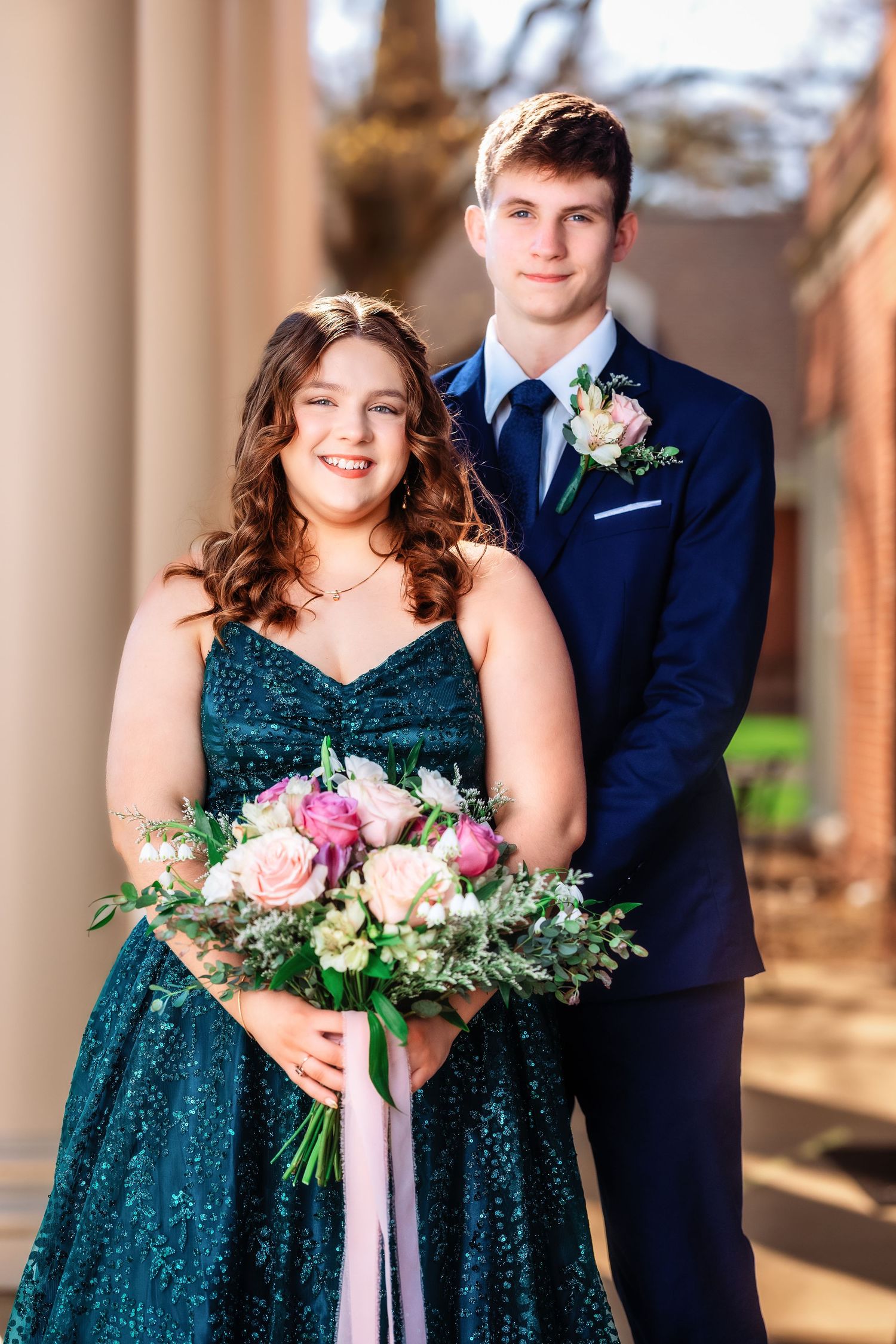 A couple in formal attire poses at prom, with her in a sparkly emerald gown holding pink flowers and him in a navy suit.