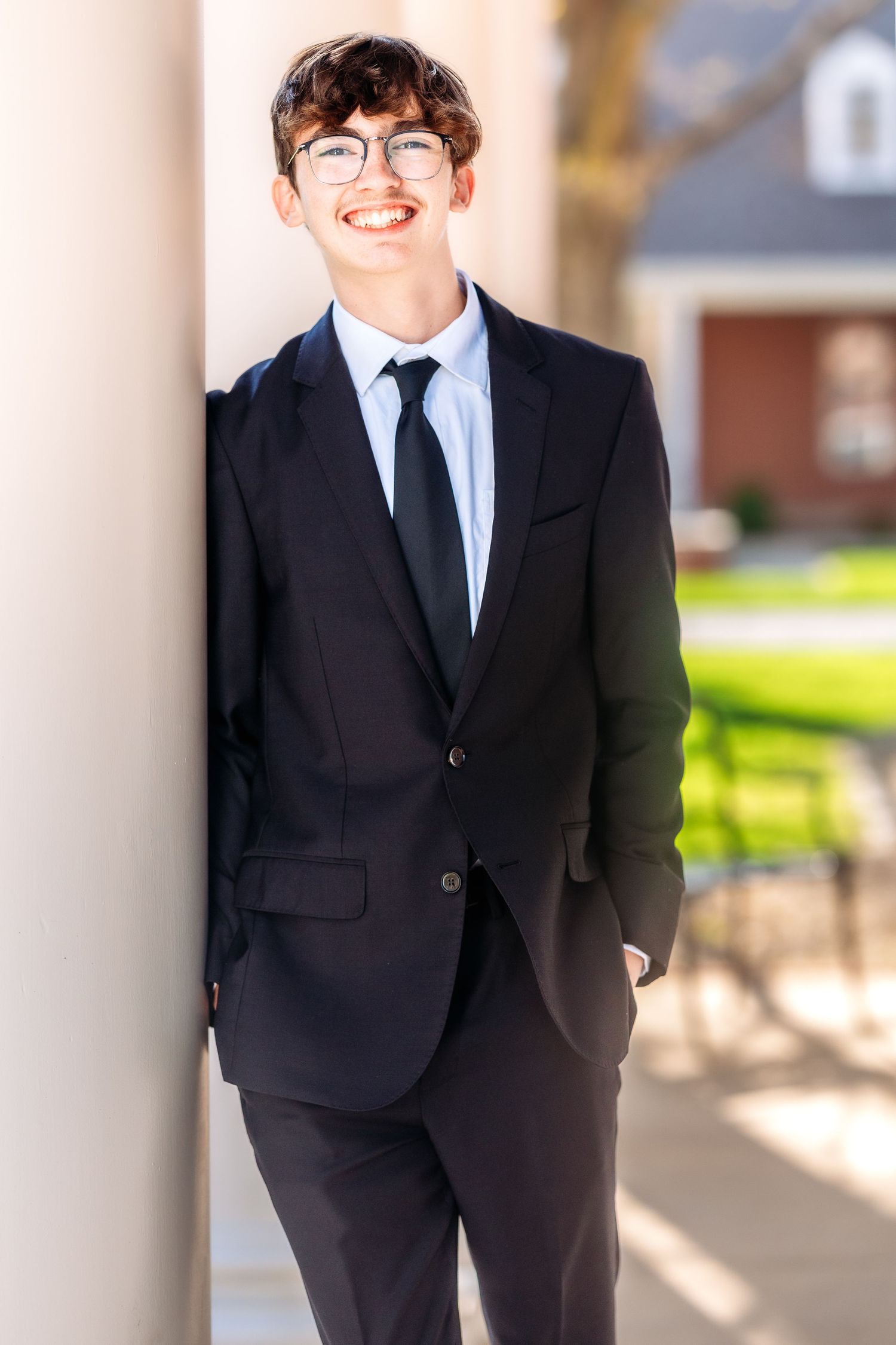 A person in a black suit and tie stands confidently against a light-colored column outdoors.