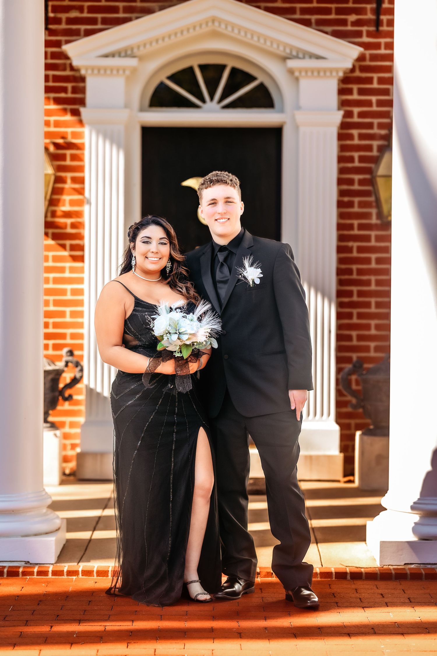 A couple in formal attire poses on brick steps of a historic building with white columns and an arched doorway.