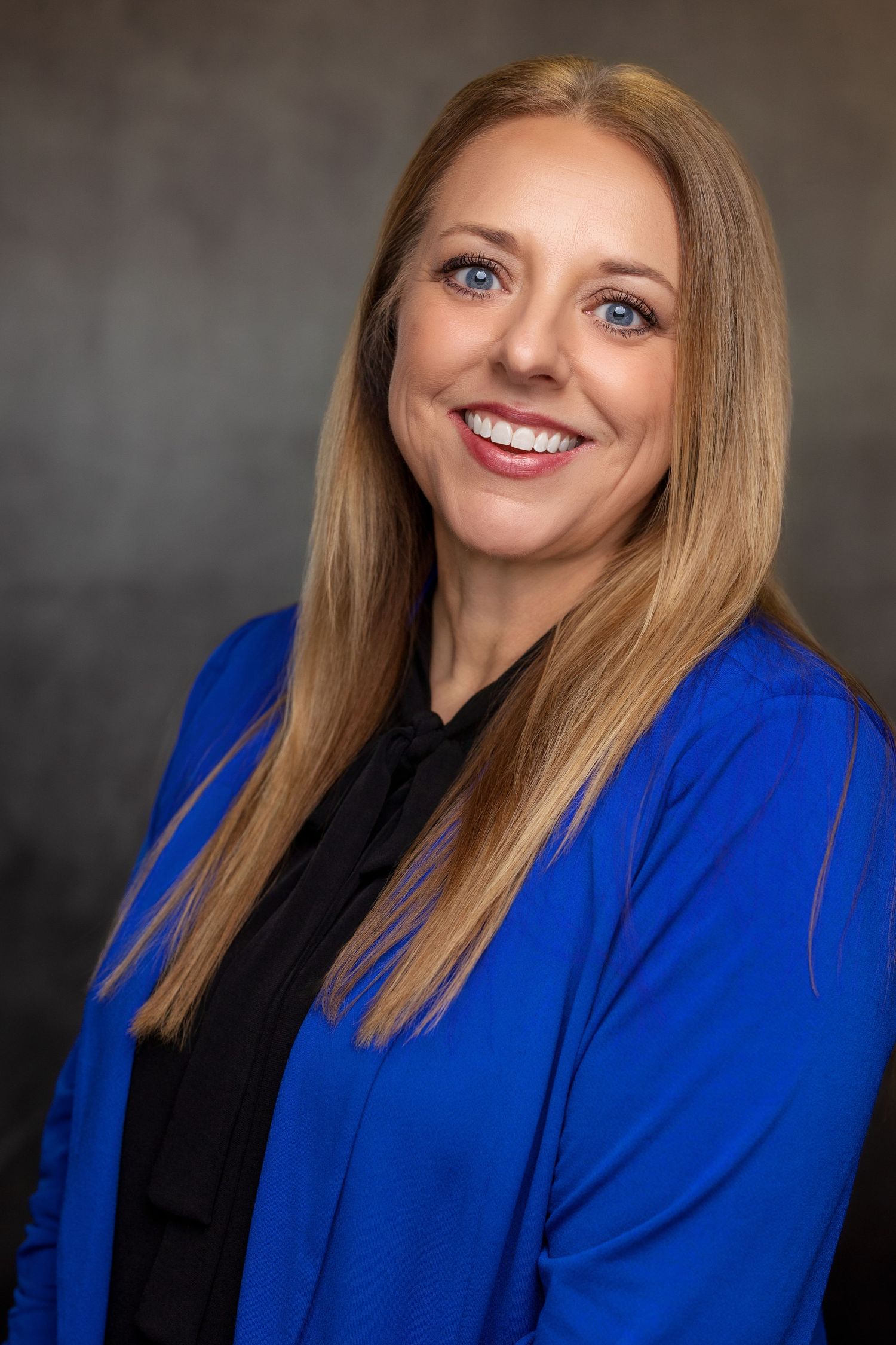 Professional headshot of person wearing royal blue blazer and black top against neutral background.