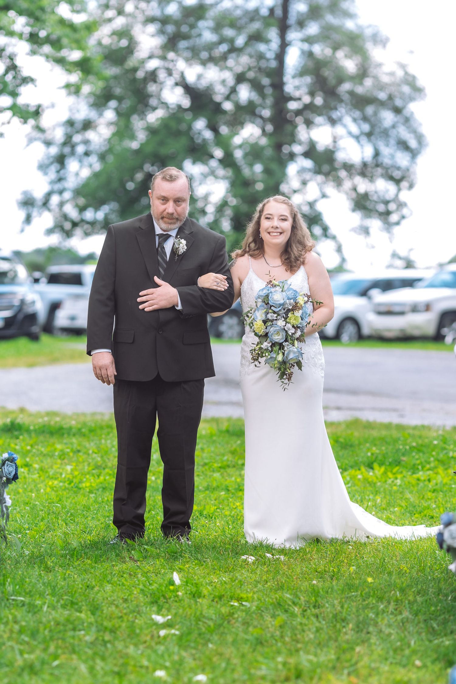 A joyful bride in white gown walks arm in arm with escort on grassy lawn with parked cars in background.