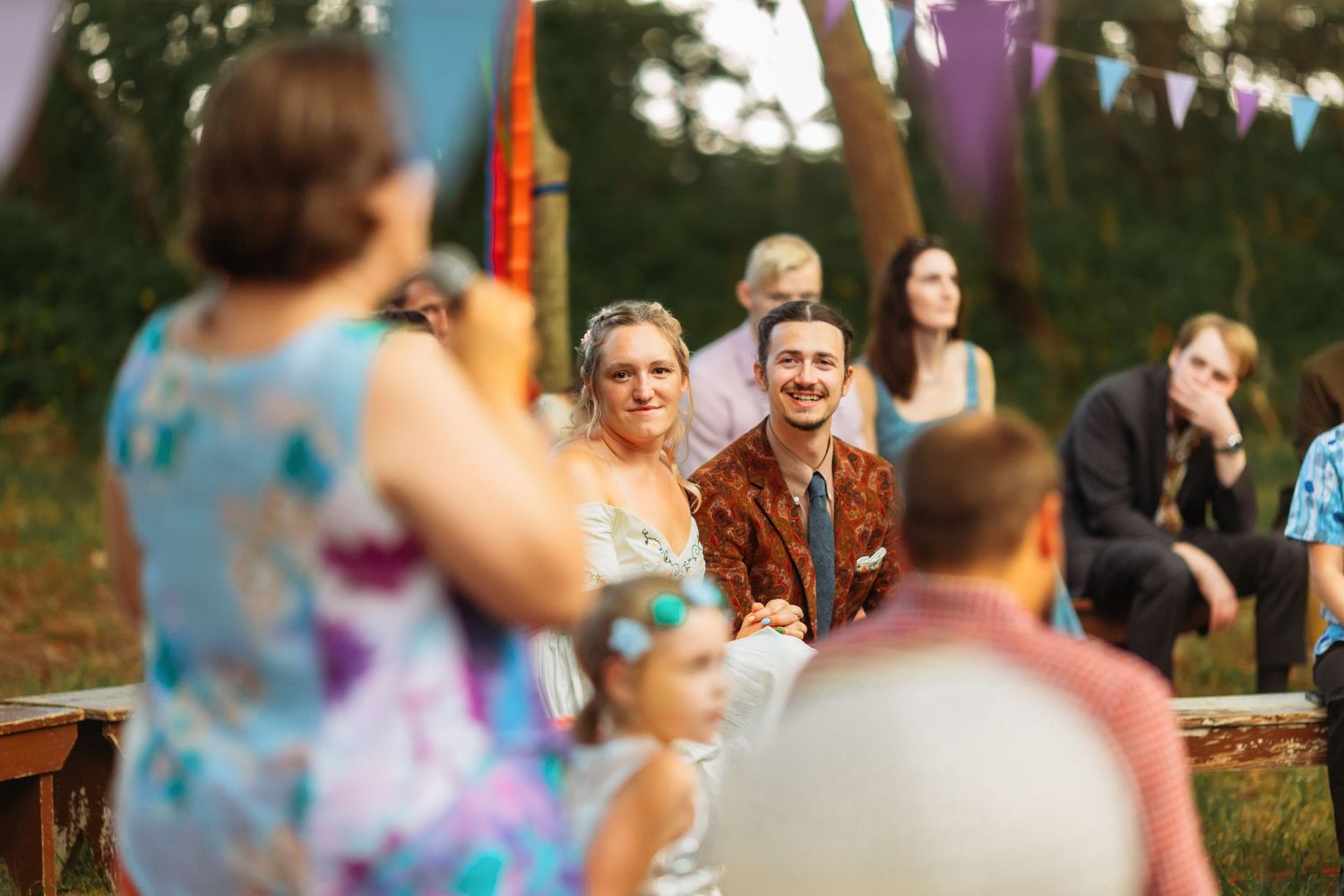 Wedding guests seated on wooden benches at an outdoor ceremony with colorful bunting decorations.
