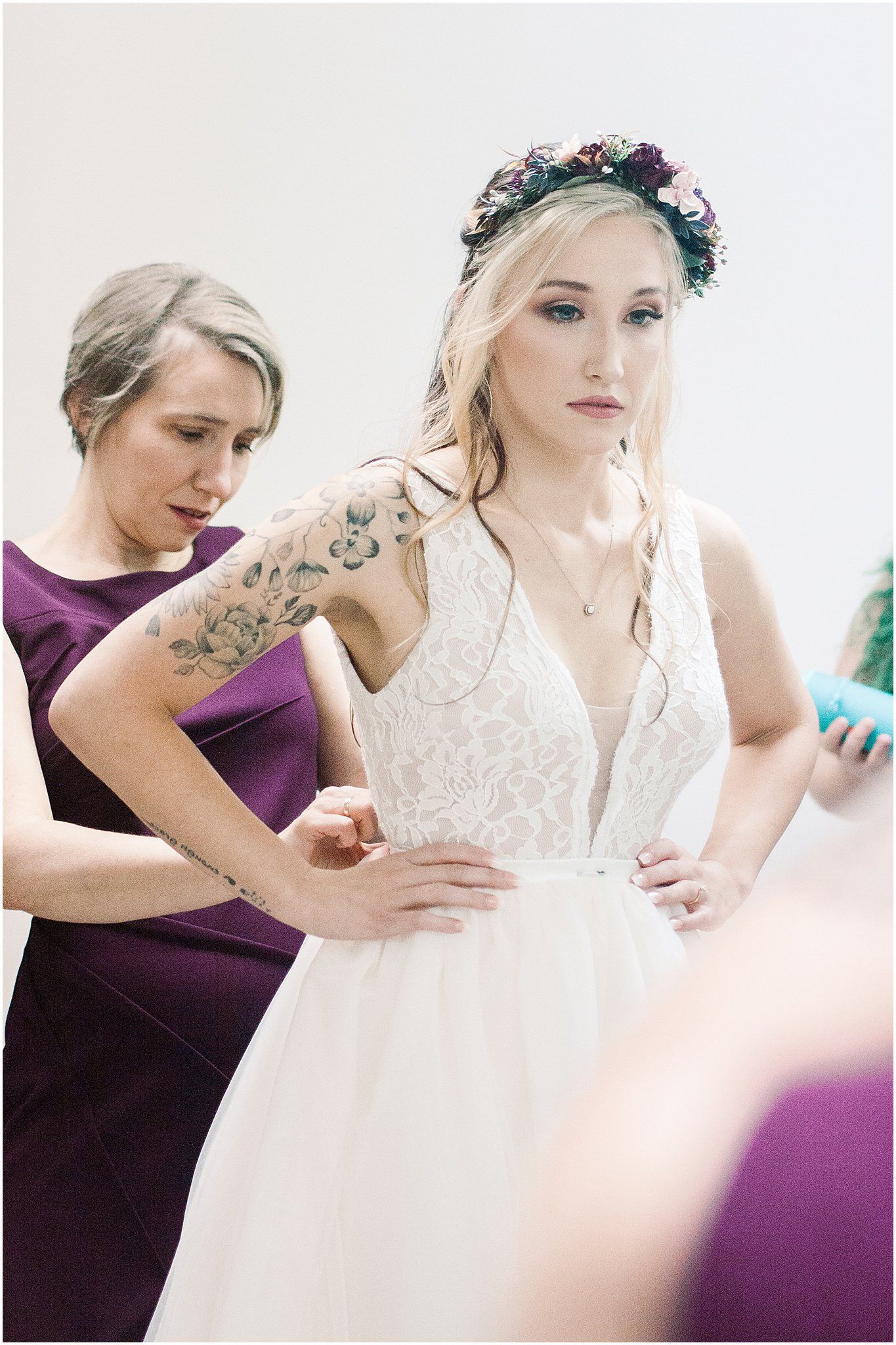 Bride in lace wedding dress with floral crown has tattoos on arm while bridesmaid helps with fitting.