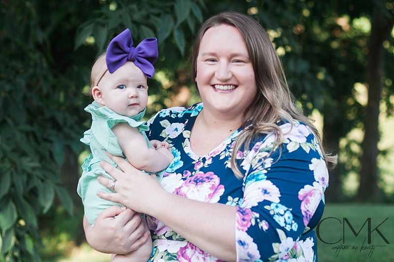A woman in floral dress holds baby in mint outfit against green foliage background.