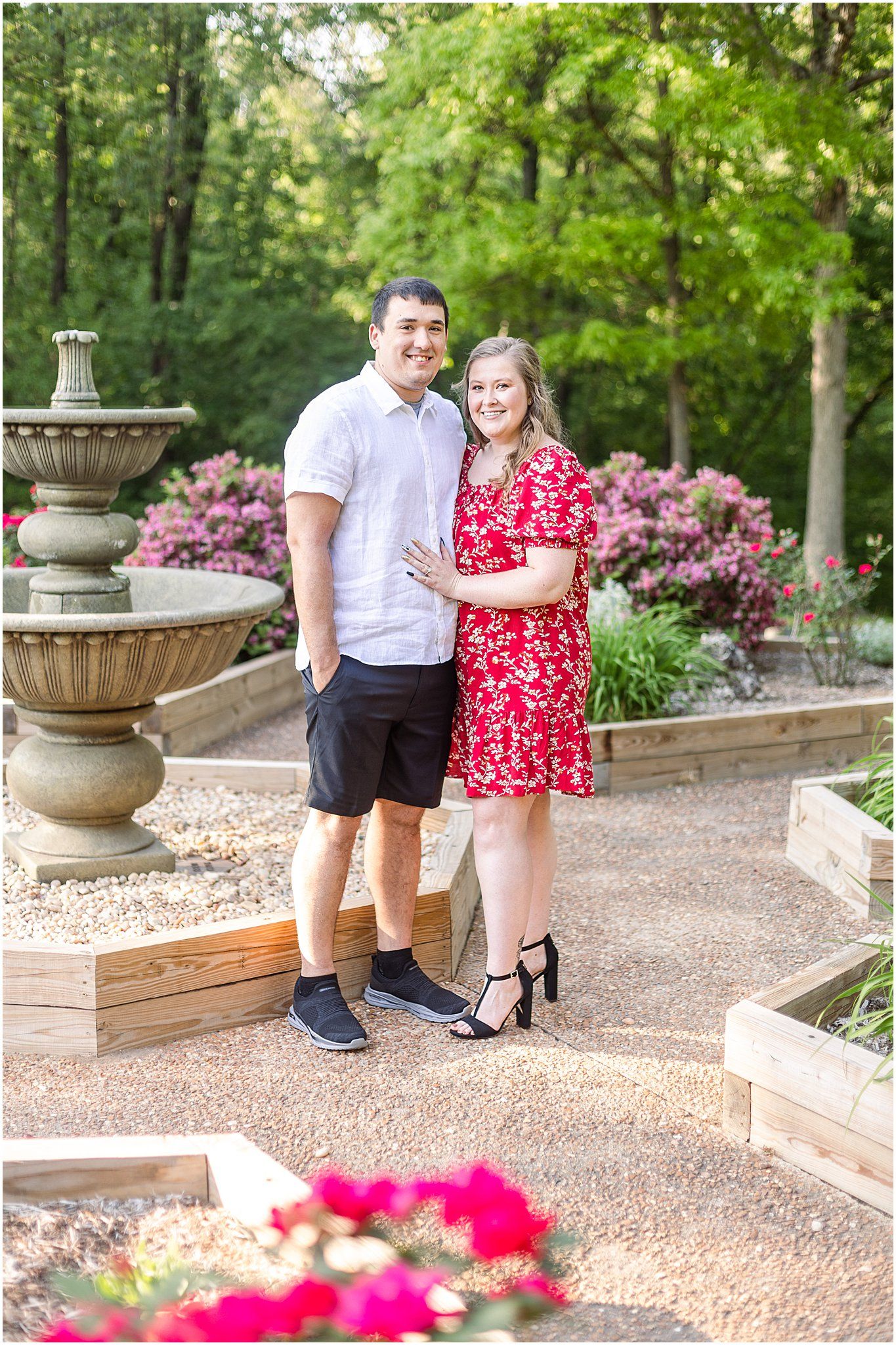 Couple posing by ornate fountain surrounded by pink flowers and landscaped garden.