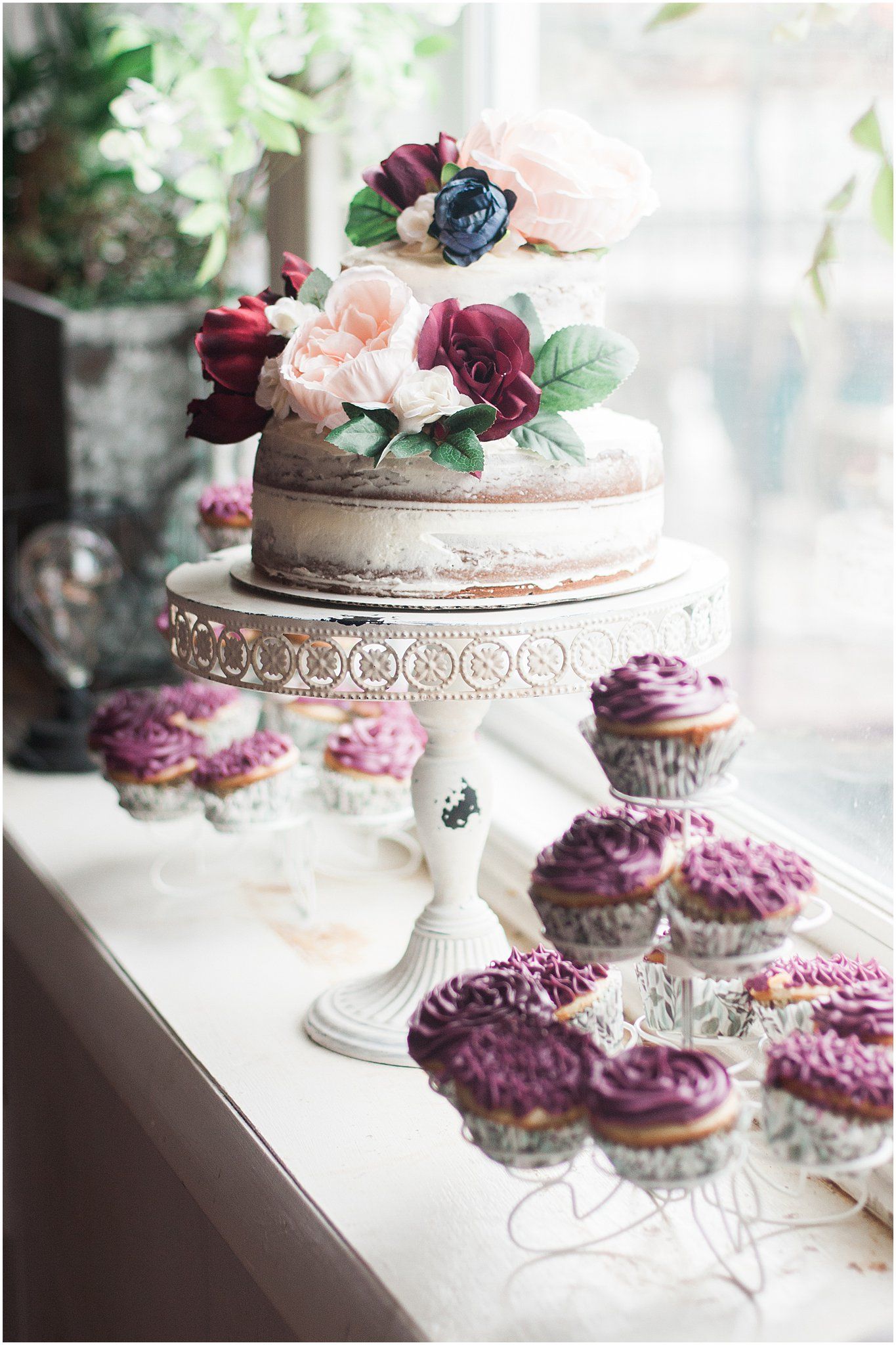 Wedding dessert display featuring a naked cake with burgundy flowers and matching cupcakes.