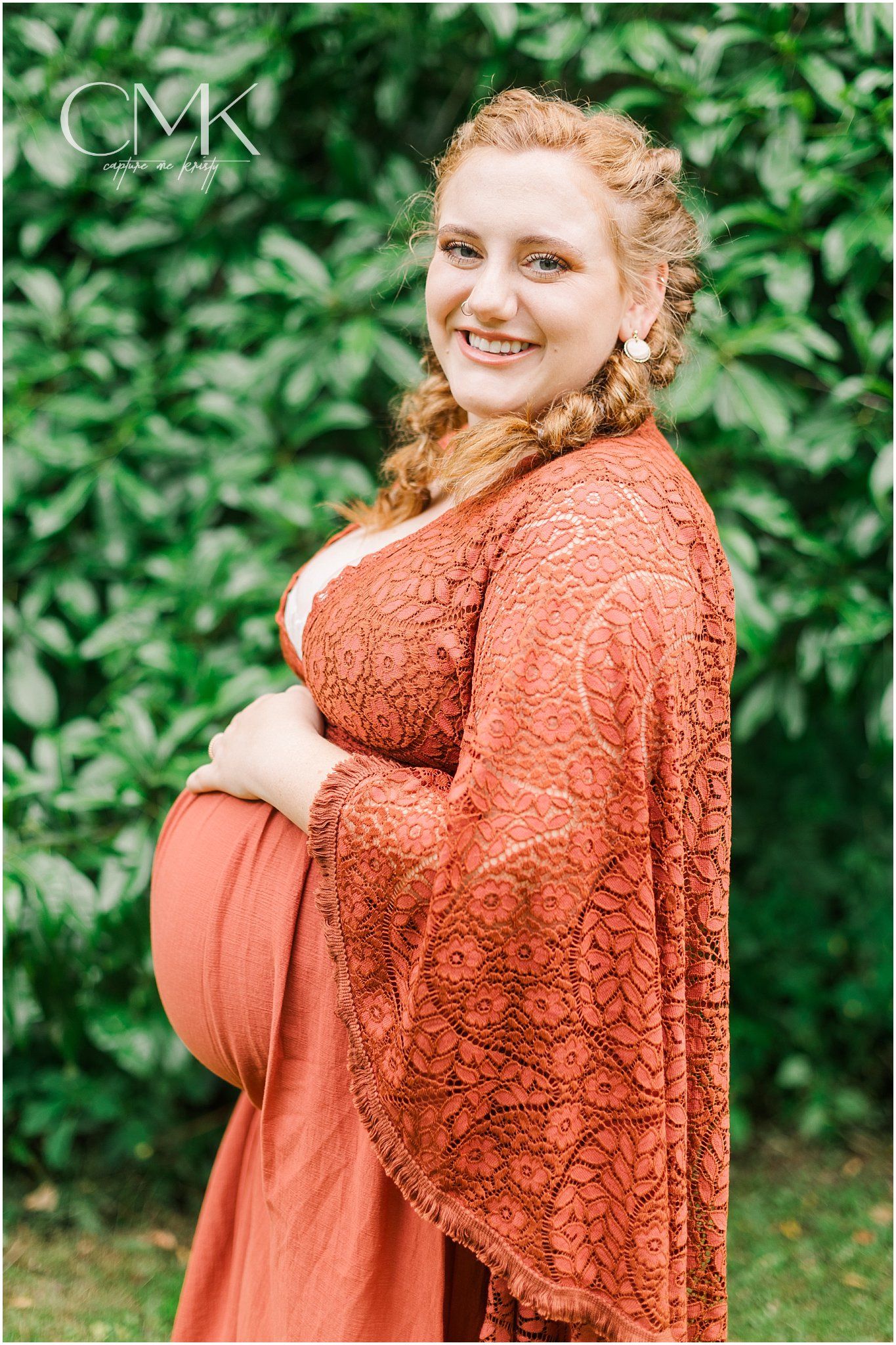 Maternity portrait in flowing coral lace dress against lush green garden backdrop.