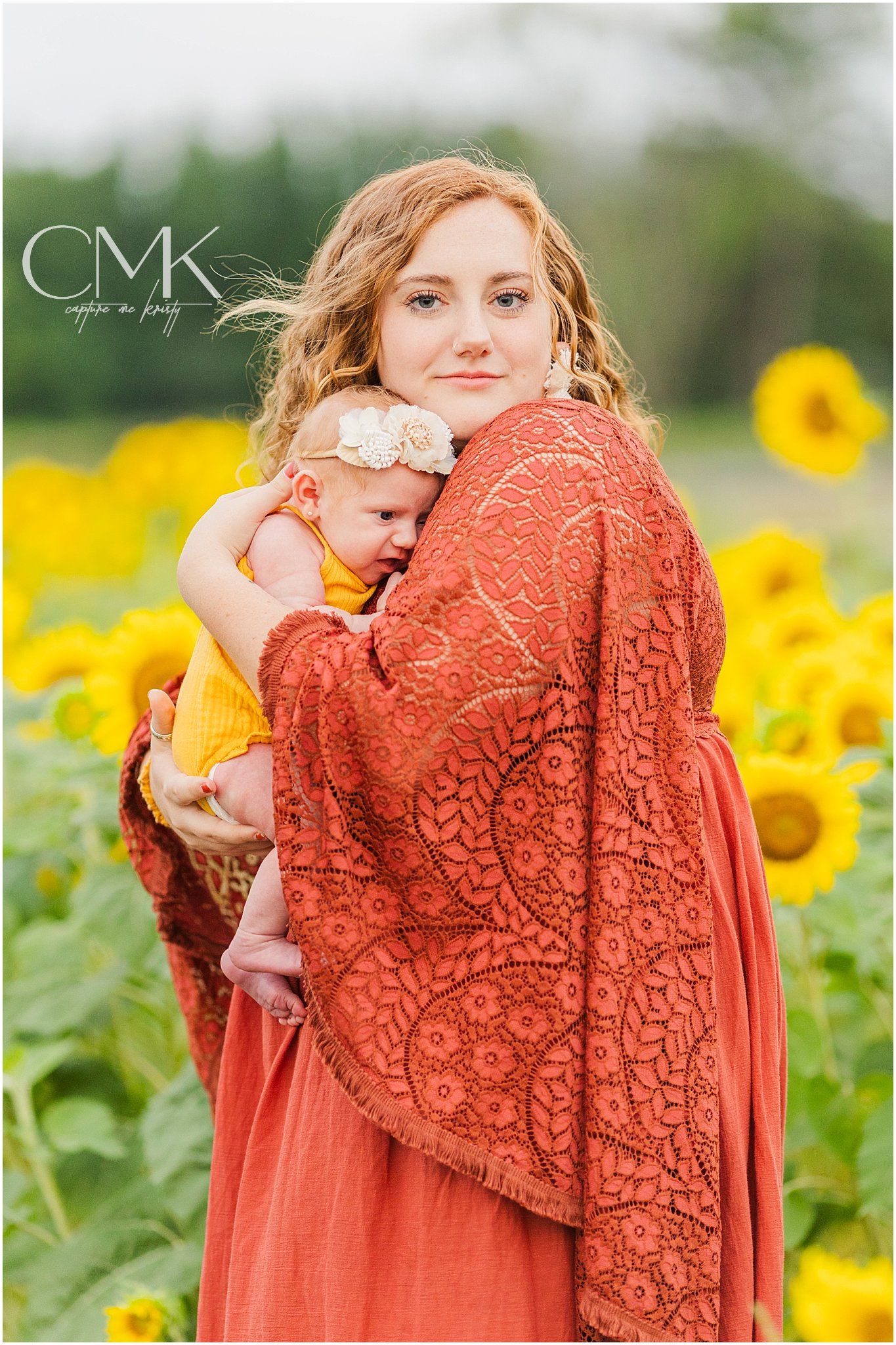 Woman in rust lace dress holds baby against backdrop of blooming sunflowers.