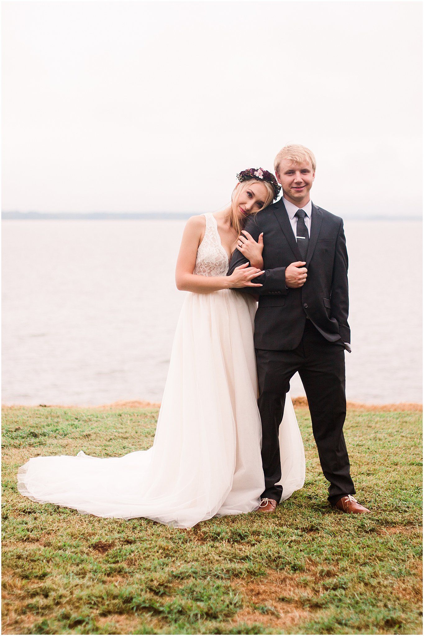 Newlyweds pose by a waterfront, with bride showing off her ring and wearing a flowing white gown.