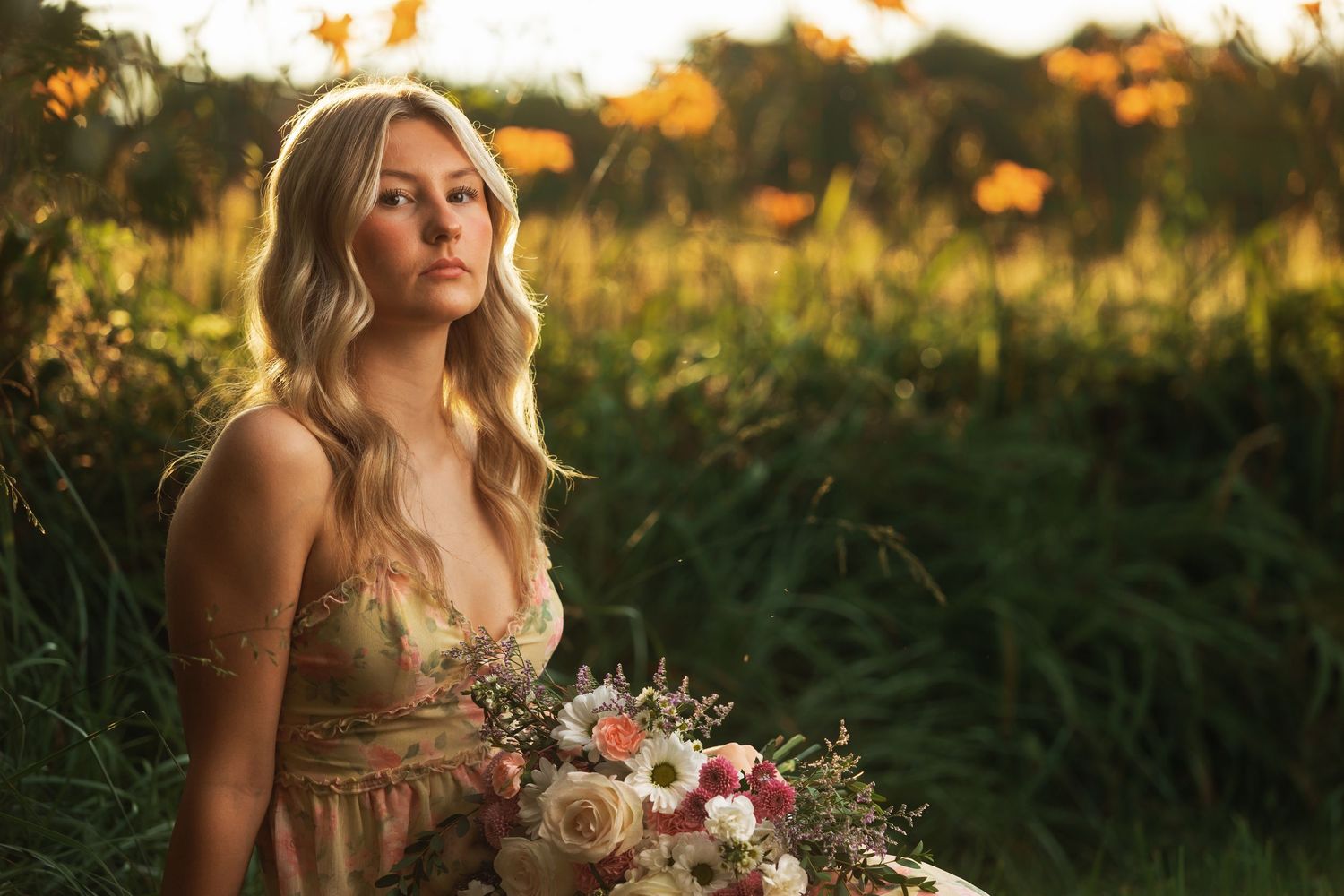 Young woman in golden dress holding floral bouquet sitting in lush green field during golden hour sunset.