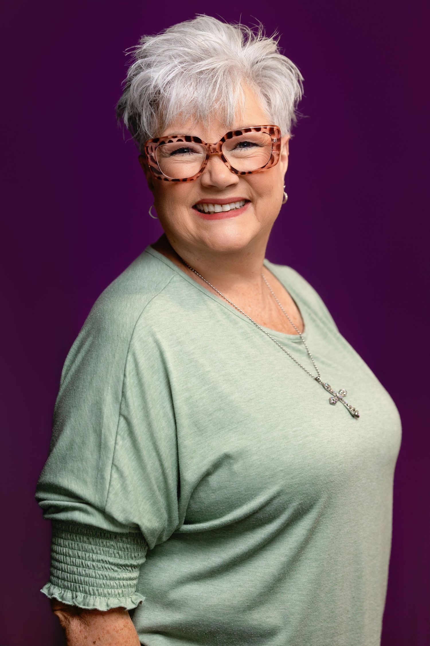 Smiling older woman with short white hair, tortoiseshell glasses, green top, and cross necklace on purple background.