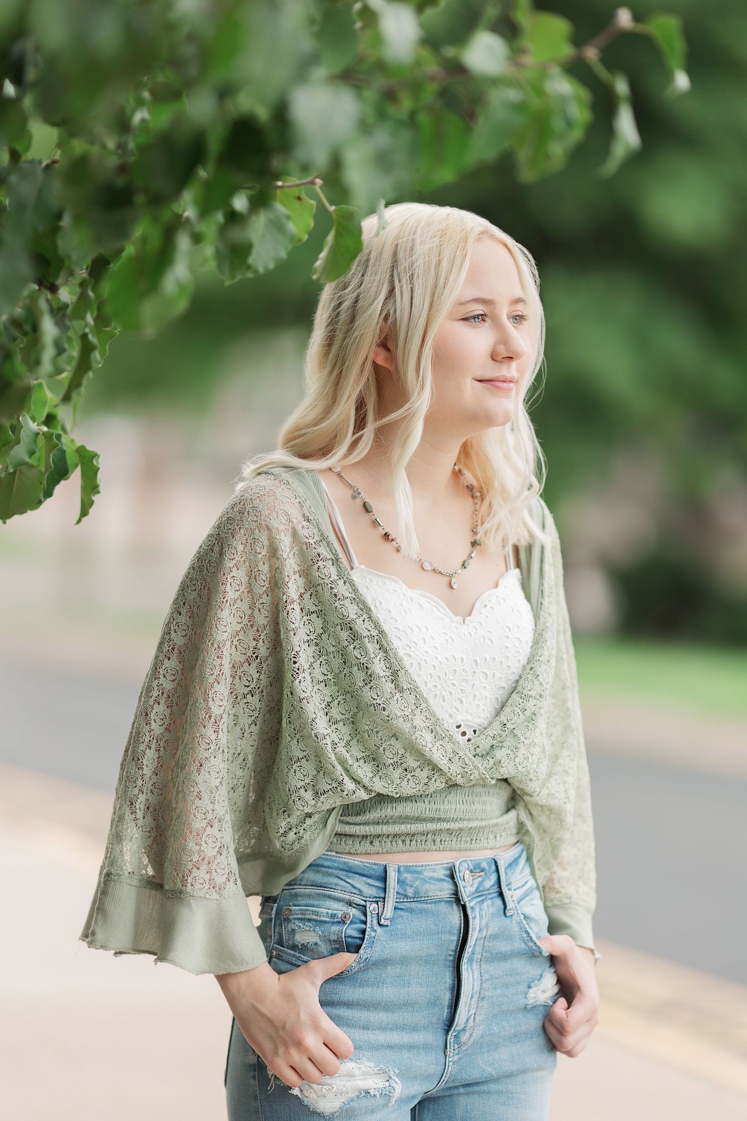 Woman in sage green lace top and jeans poses against blurred natural background.
