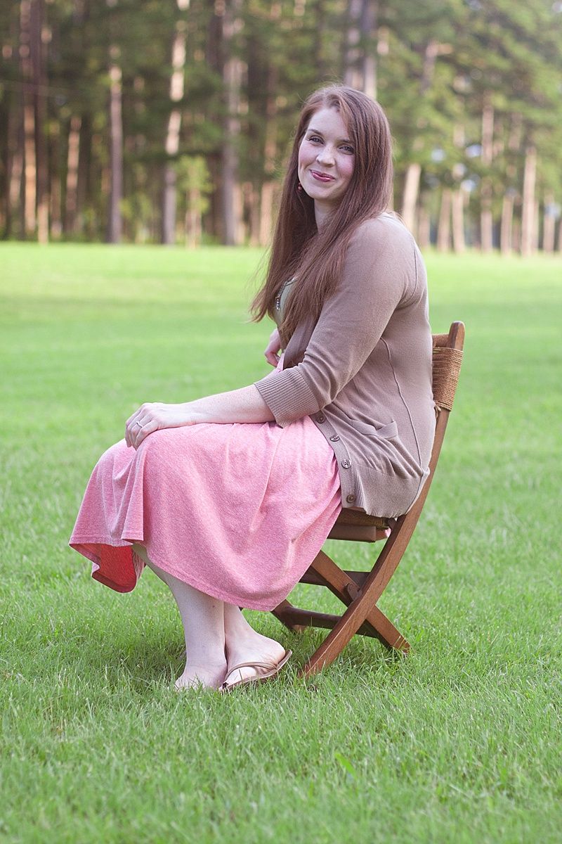 Woman in brown sweater and pink skirt sits on wooden chair in peaceful garden setting.
